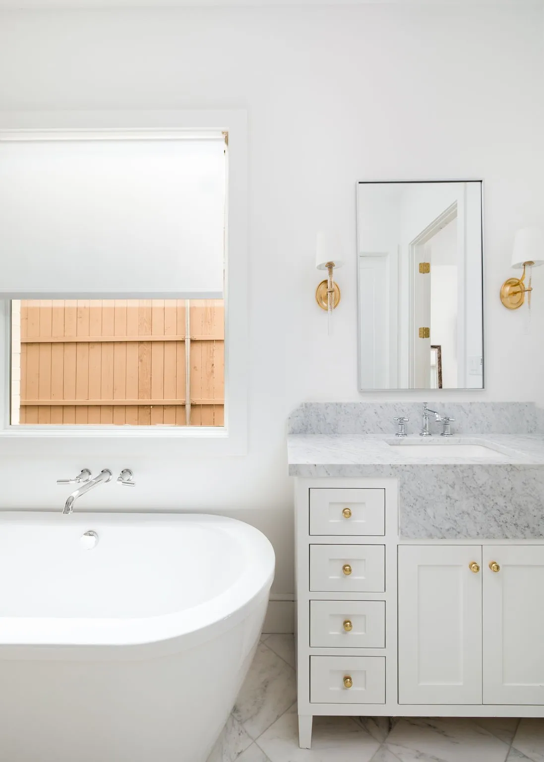 Freestanding soaking tub with wall-mounted faucet, single vanity with white cabinetry, stone countertop, undermount sink, and wall-mounted sconces