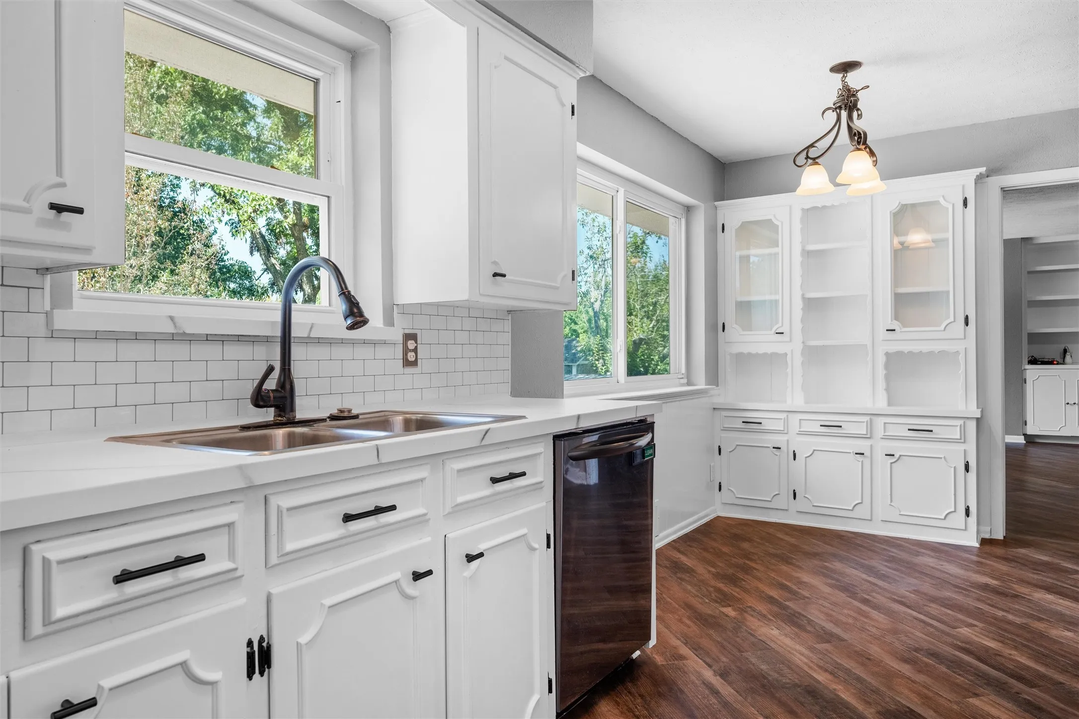 White shaker-style cabinetry with dark hardware, a double basin sink, subway tile backsplash, and wood-finish flooring