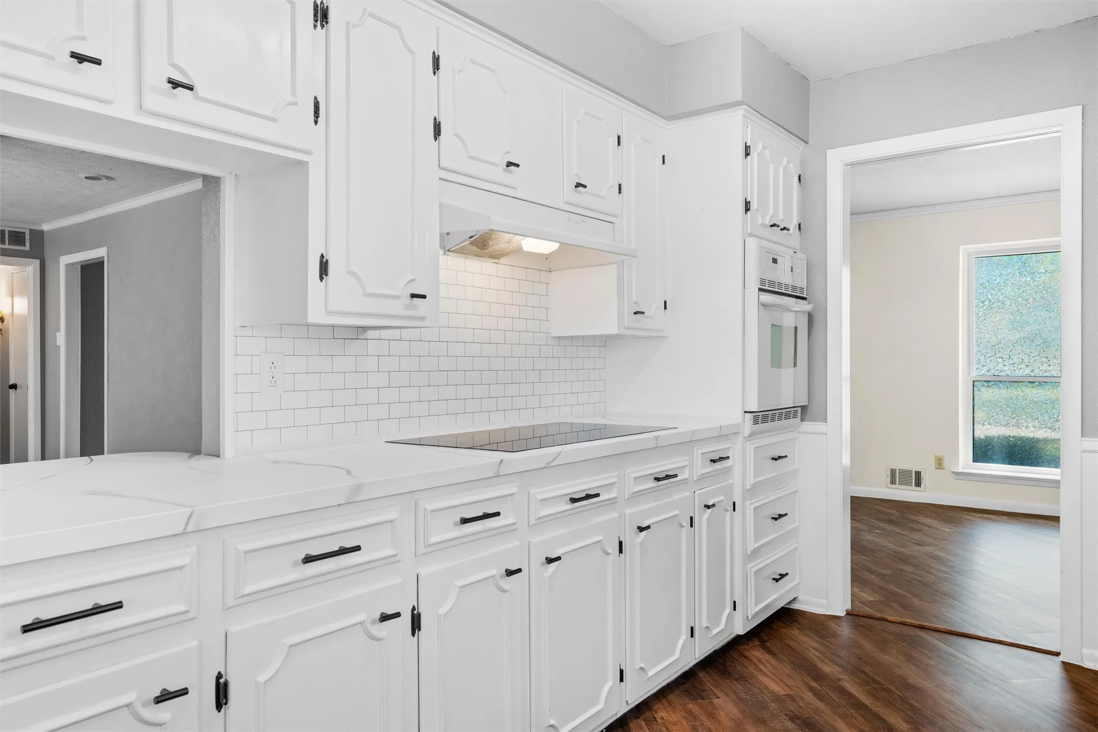 White cabinetry with black hardware, subway tile backsplash, white countertops, built-in double oven, and wood-finish flooring