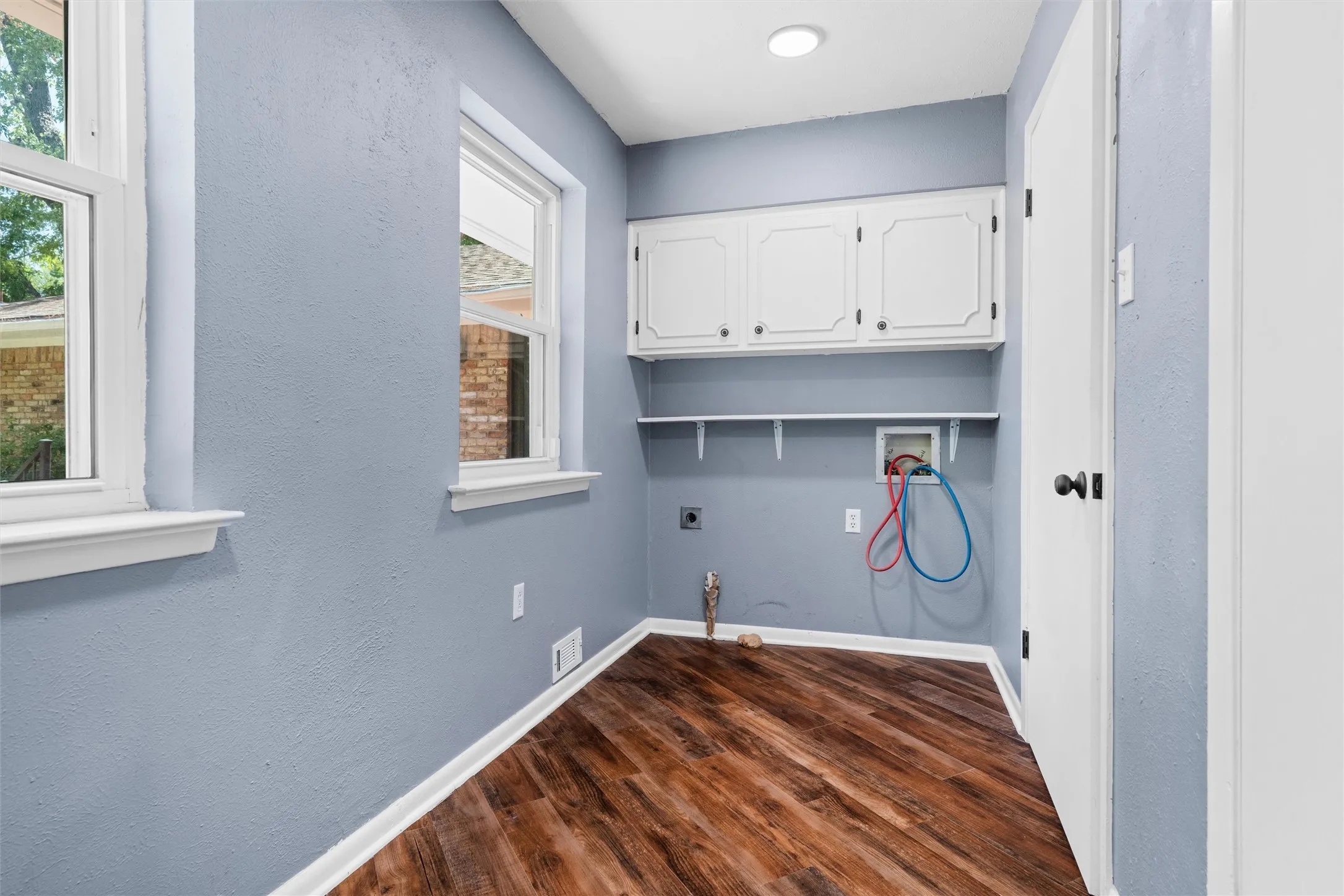 Laundry area featuring wood-finish flooring, two windows, wall-mounted shelving, and upper cabinetry