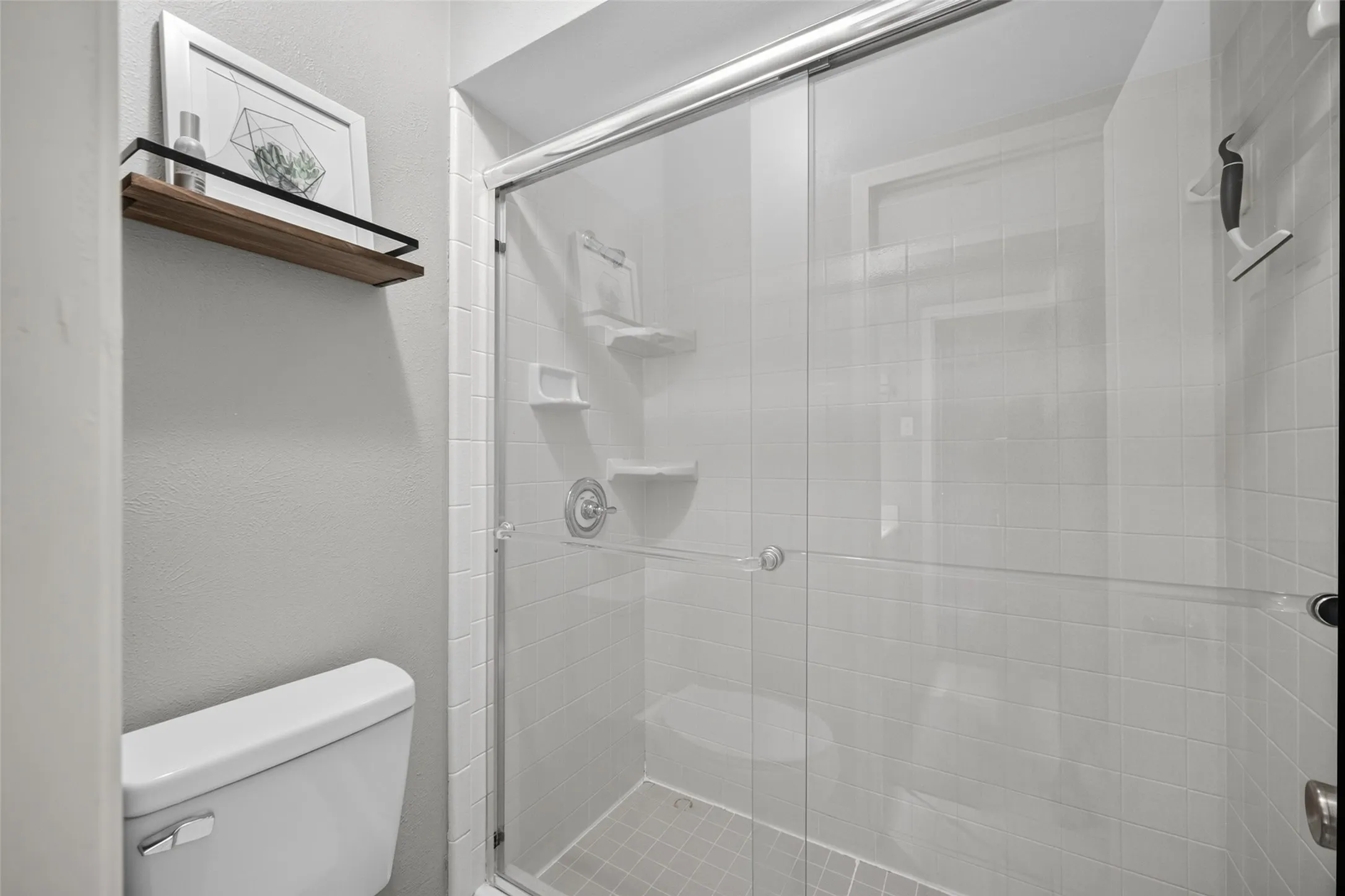 Shower with sliding glass enclosure, white square tile surround, recessed shelving, and a toilet