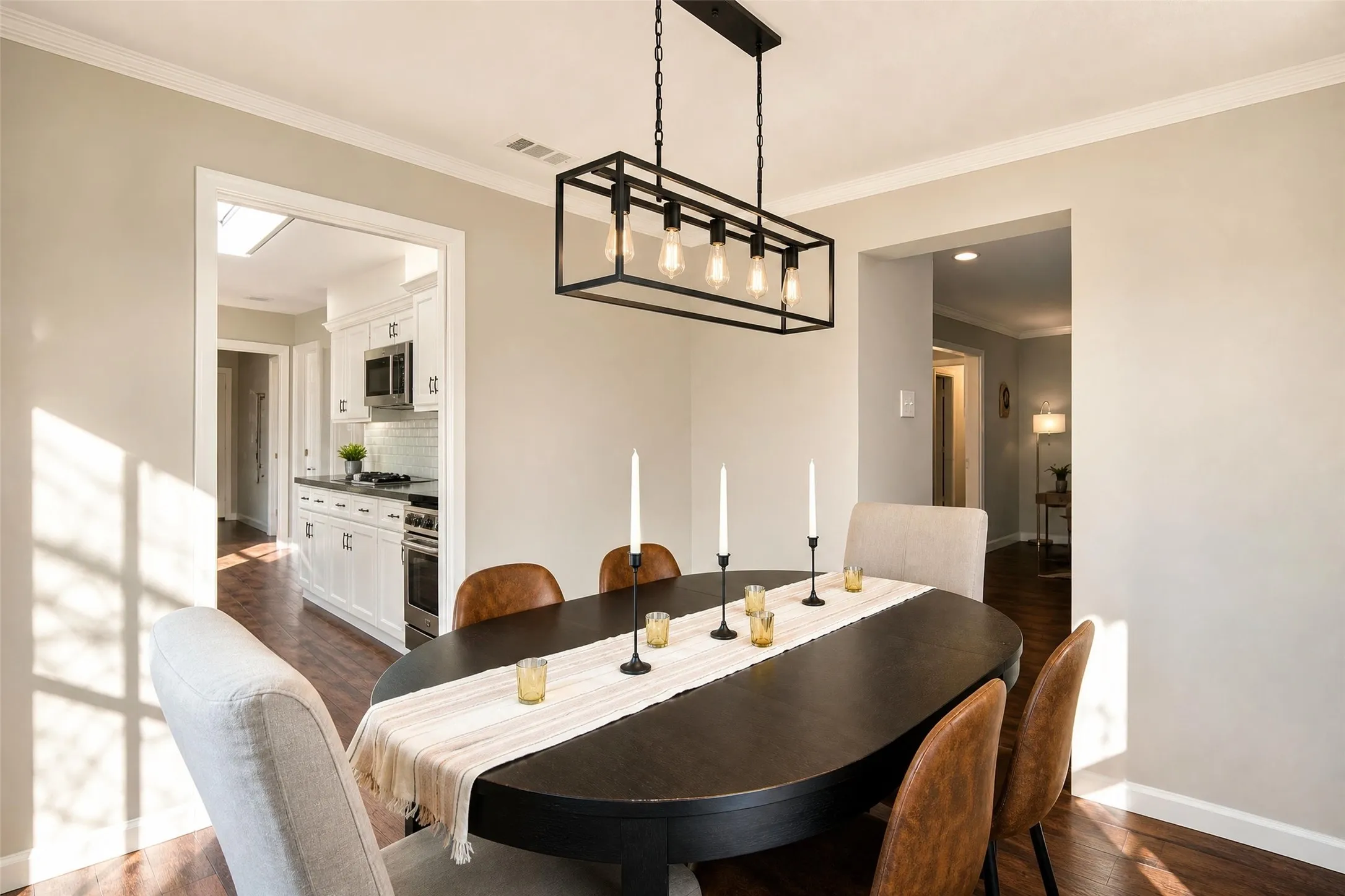 Dining area with wood-finish flooring, a rectangular linear chandelier, and an open doorway to a kitchen featuring white cabinetry and stainless steel appliances