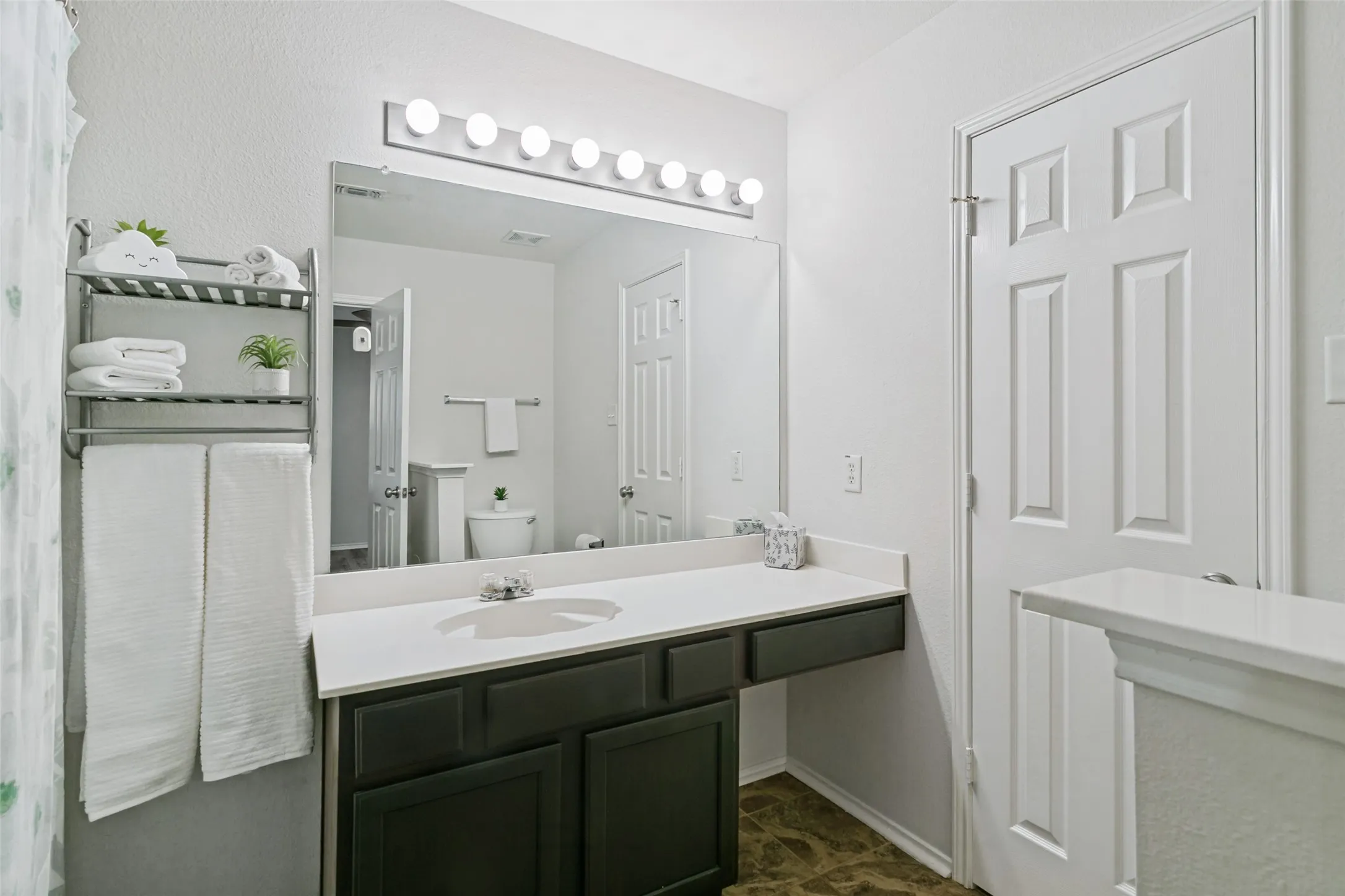 White paneled door, dark wood-finish vanity with white countertop, chrome single-handle faucet, illuminated mirror, and a wall-mounted shelving unit