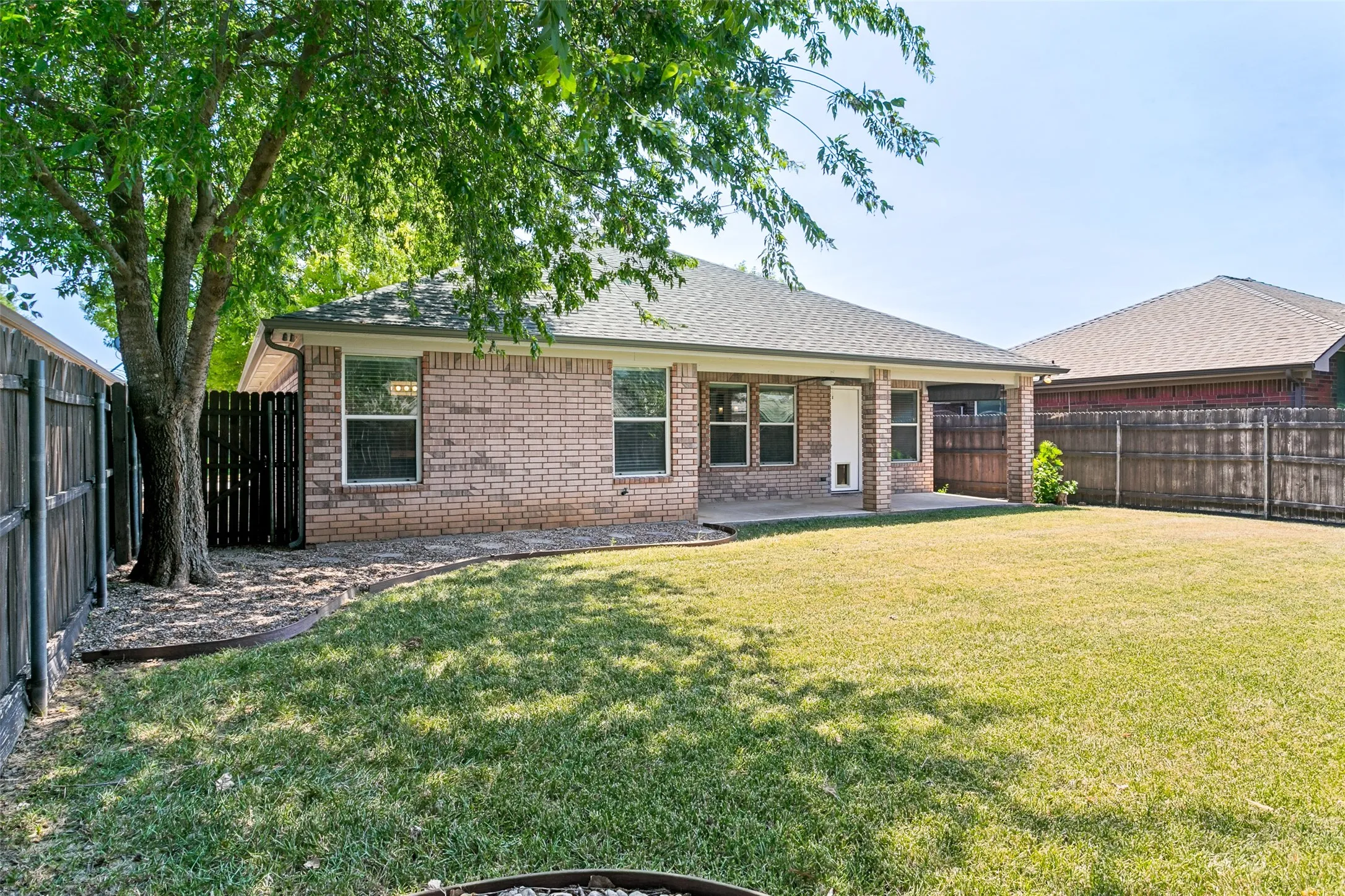 Expansive backyard featuring a manicured lawn, full privacy fencing, mature shade tree, and a covered patio