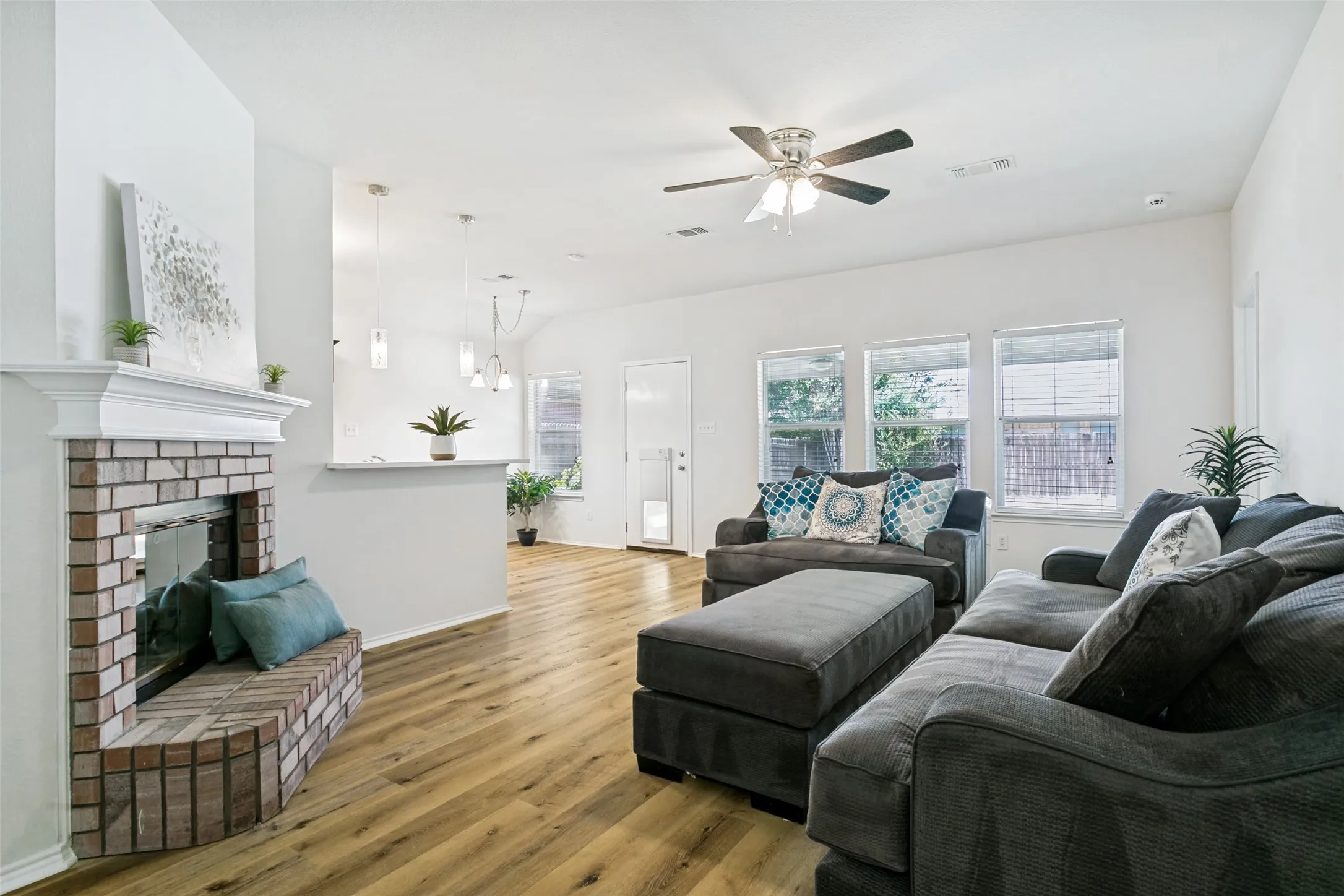 Brick fireplace with hearth, wood-finish flooring, multiple windows, ceiling fan, and pendant lighting