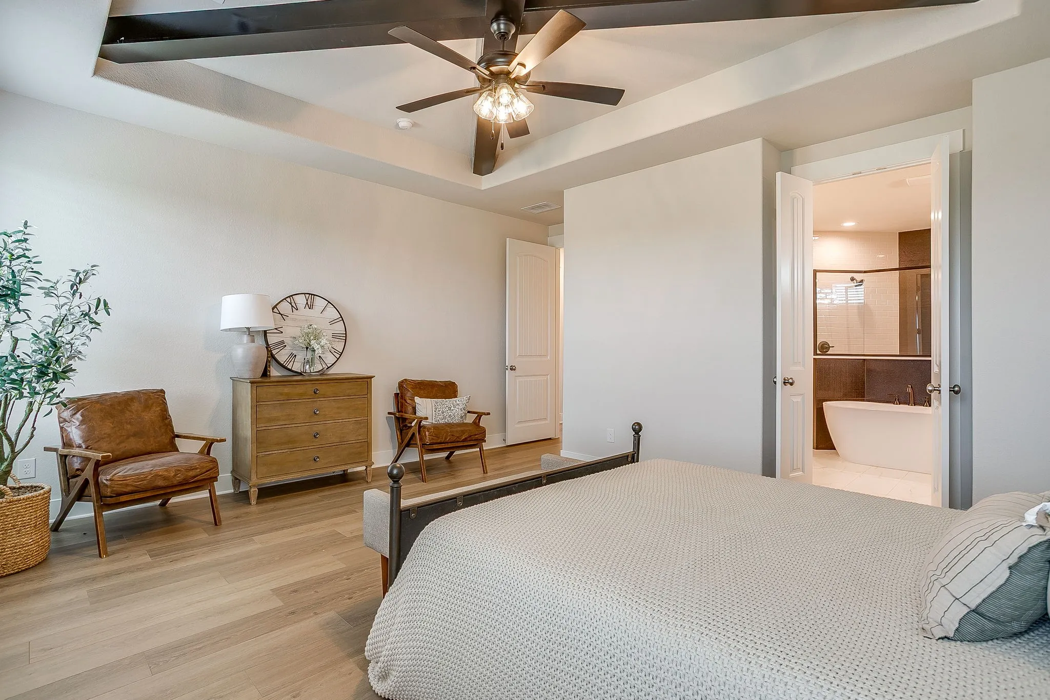 Tray ceiling with exposed beams, wood-finish flooring, and an attached full bathroom featuring a freestanding soaking tub and glass-enclosed shower