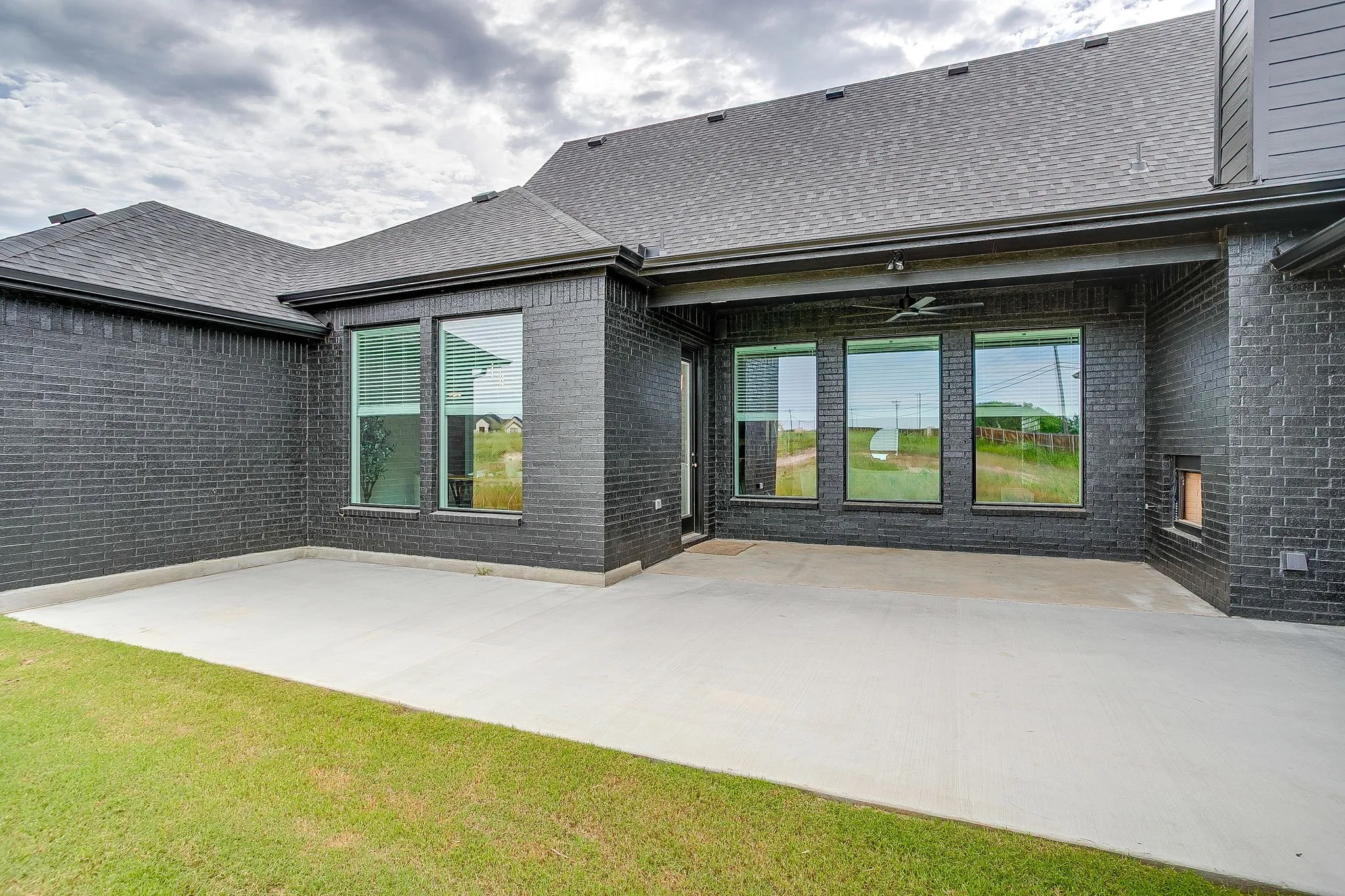 Exterior featuring dark brick facade, a covered patio with an installed ceiling fan, a concrete patio surface, and multiple large windows