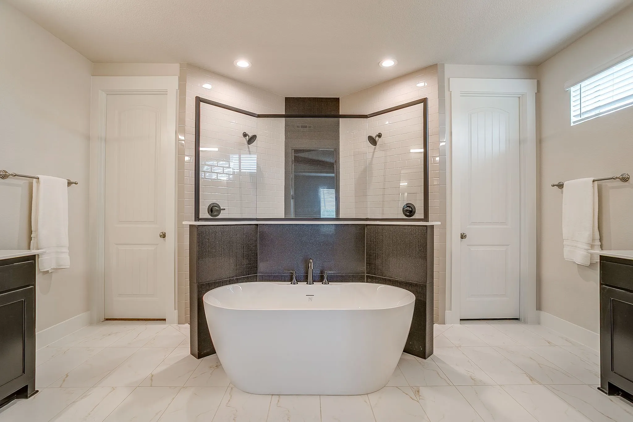 Freestanding soaking tub, dual showers with black hardware, white subway tile shower surrounds, white marble-look flooring, and recessed lighting