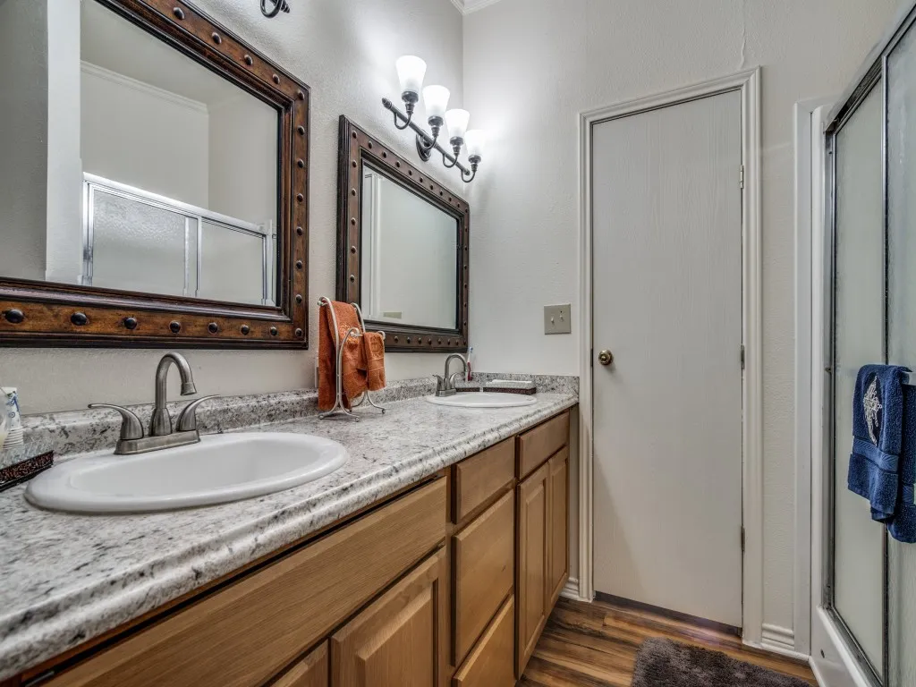 Dual vanity with white oval sinks and brushed nickel faucets, granite-look countertop, wood-finish cabinetry, wood-look flooring, and a glass-enclosed shower