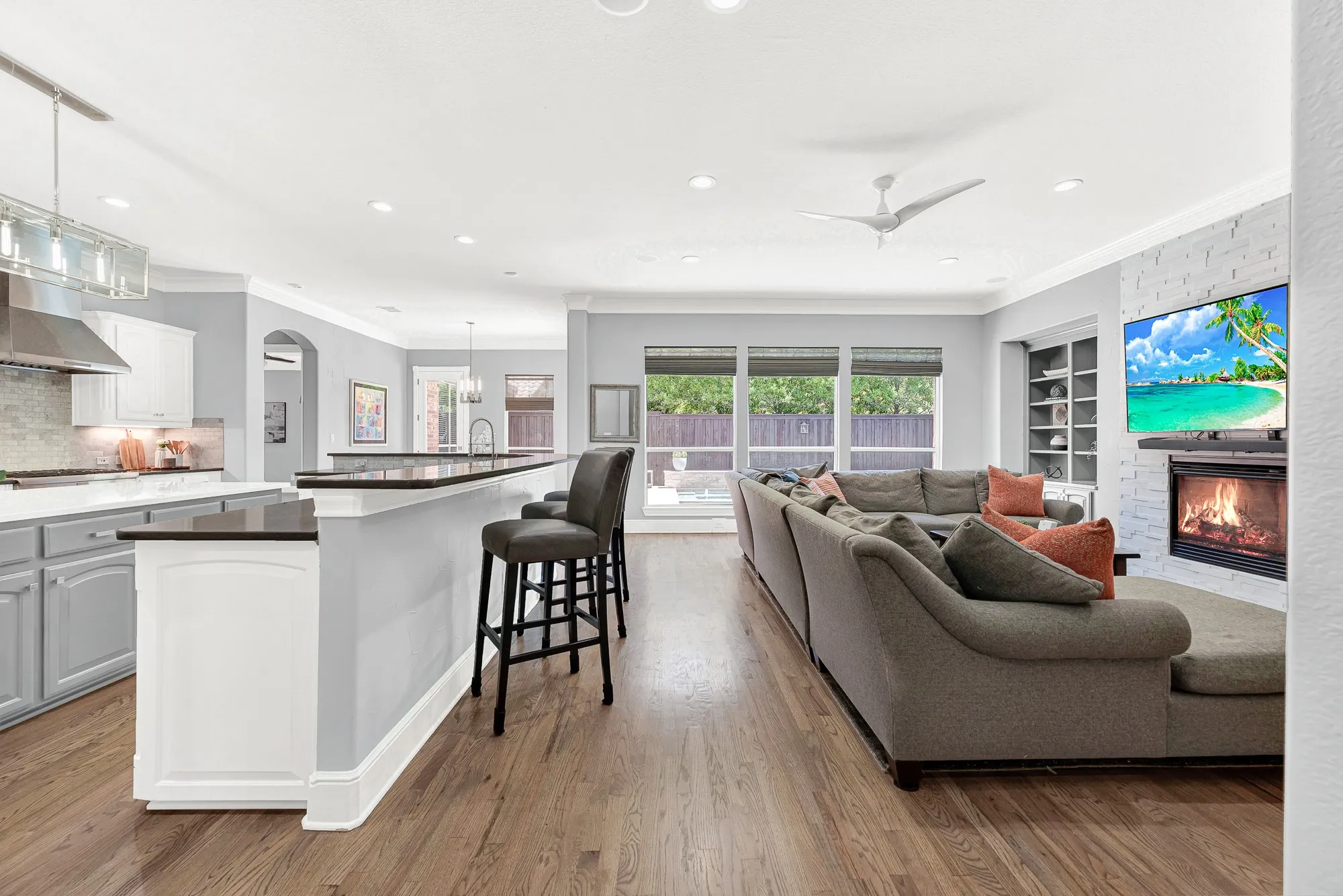Warm wood floors flow past the raised breakfast bar into a light-filled living area featuring a stacked-stone fireplace and rear windows opening to the pool. Enjoy effortless entertaining as friends mingle between the kitchen counter and sunny outdoor patio views.