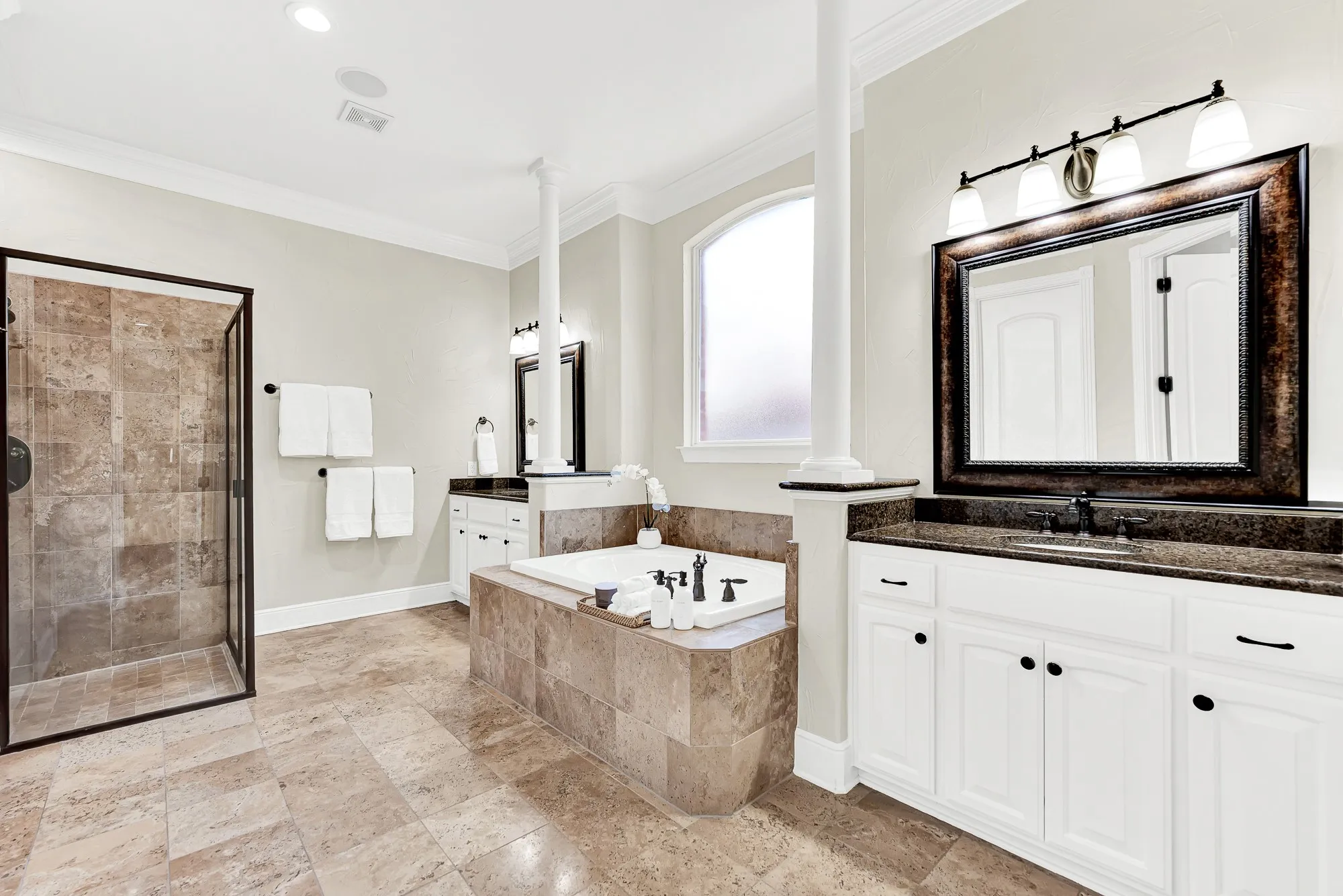 Rich tile flooring surrounds dual vanities, a deep soaking tub, and a glass walk-in shower in this luxurious primary bathroom. Stream gentle morning music through the whole-home audio system while preparing for your day in complete spa-like luxury.