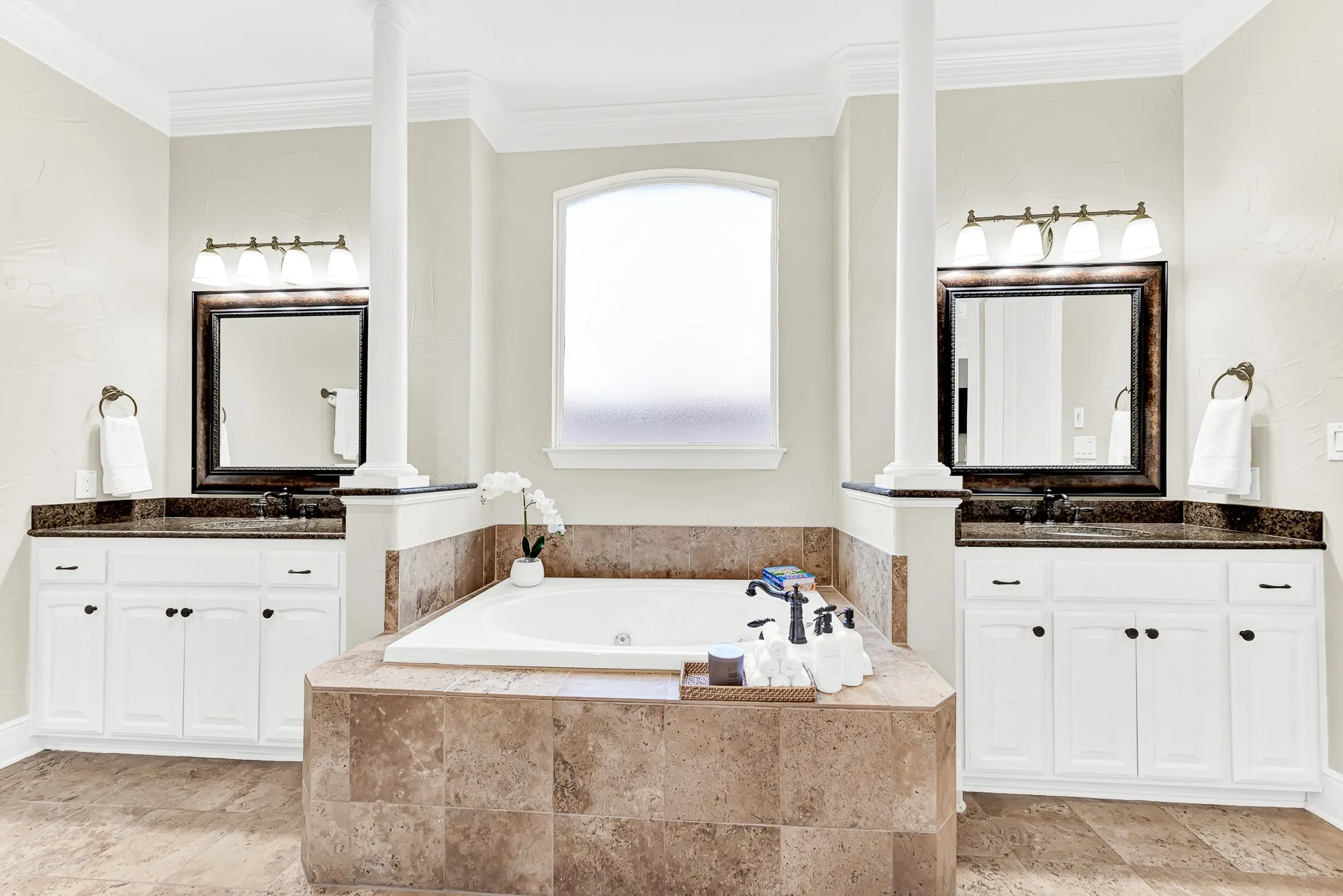 An arched frosted window lights the deep central soaking tub, flanked symmetrically by white vanities and elegant support columns. Treat yourself to a long evening soak and transform daily bath routines into pure relaxation.