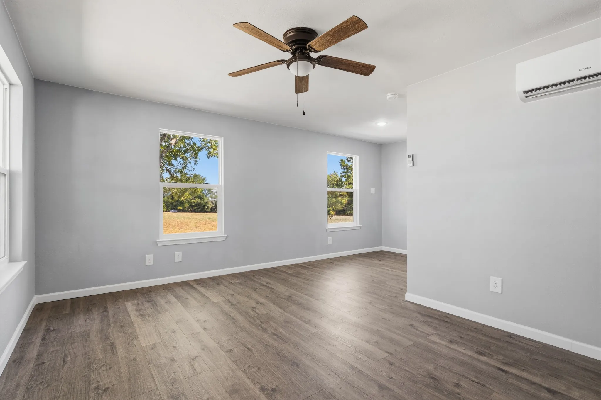 Spacious room featuring wood-finish flooring, multiple windows, a ceiling fan, and a wall-mounted climate control unit