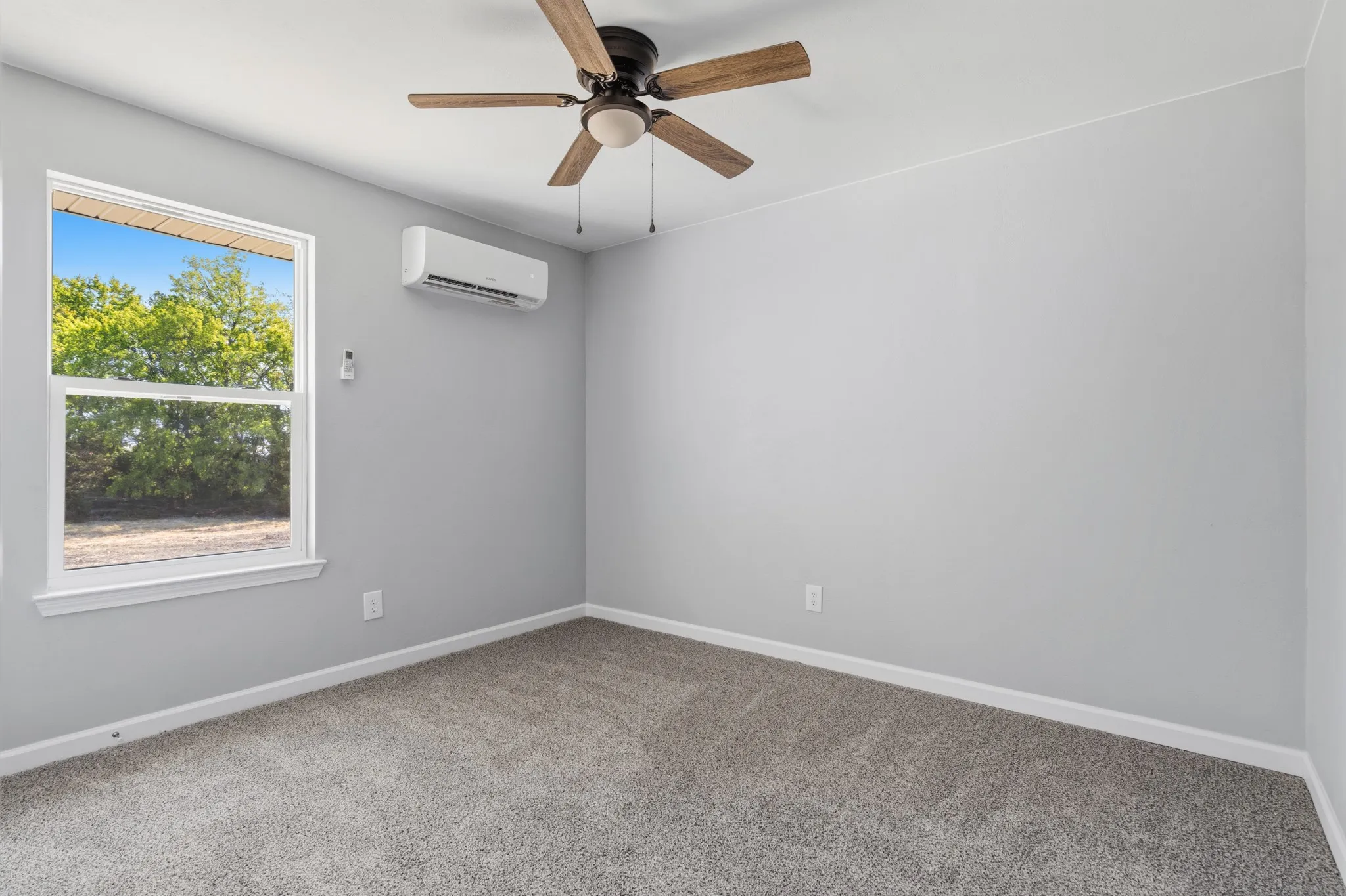 Light gray wall finish, gray carpet flooring, ceiling fan with wood-finish blades, white window trim, and a wall-mounted climate control unit