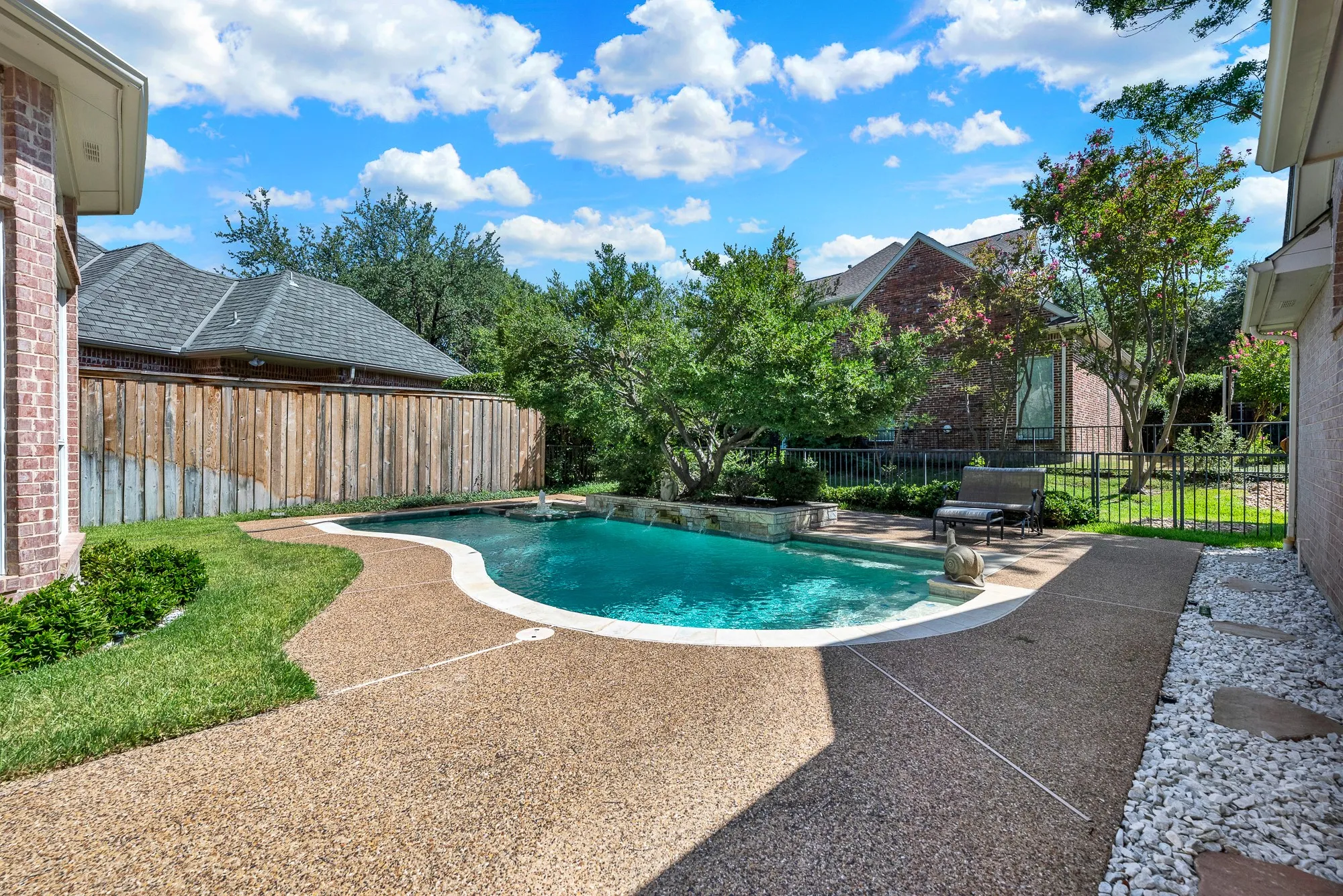 Smooth coping surrounds the gently curved pool basin, reflecting bright open skies above a surrounding privacy fence and manicured lawn accents. Picture taking a refreshing mid-day dip before cooling off on the sunny deck with an iced tea