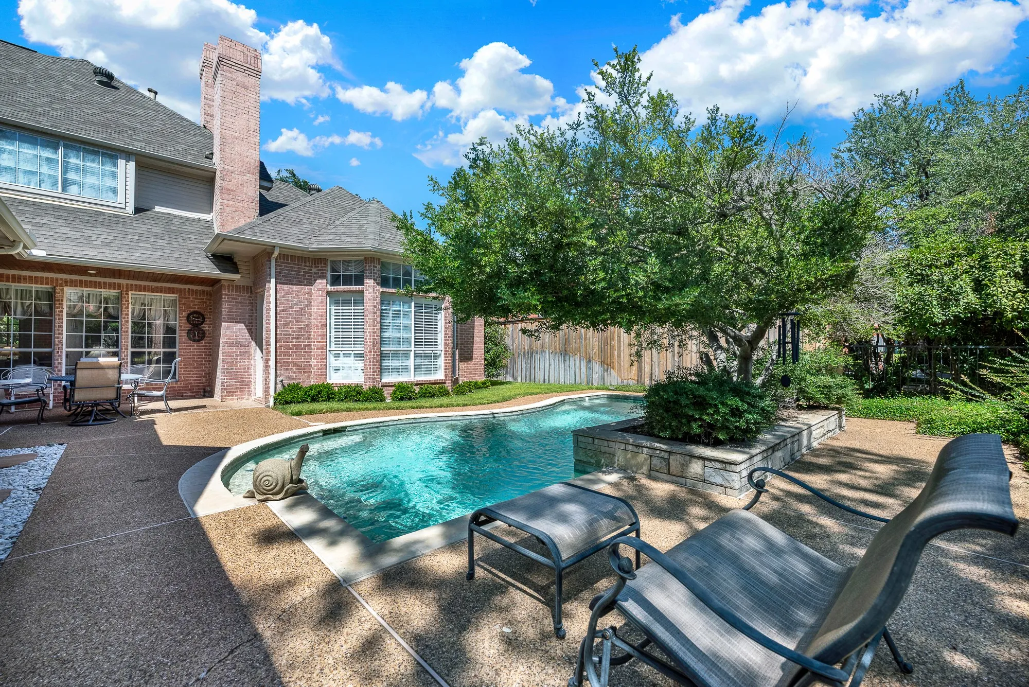 Curving aggregate decking frames a freeform swimming pool nestled under a leafy shade tree. Picture spending lazy summer afternoons lounging poolside while catching a cool breeze in the shade