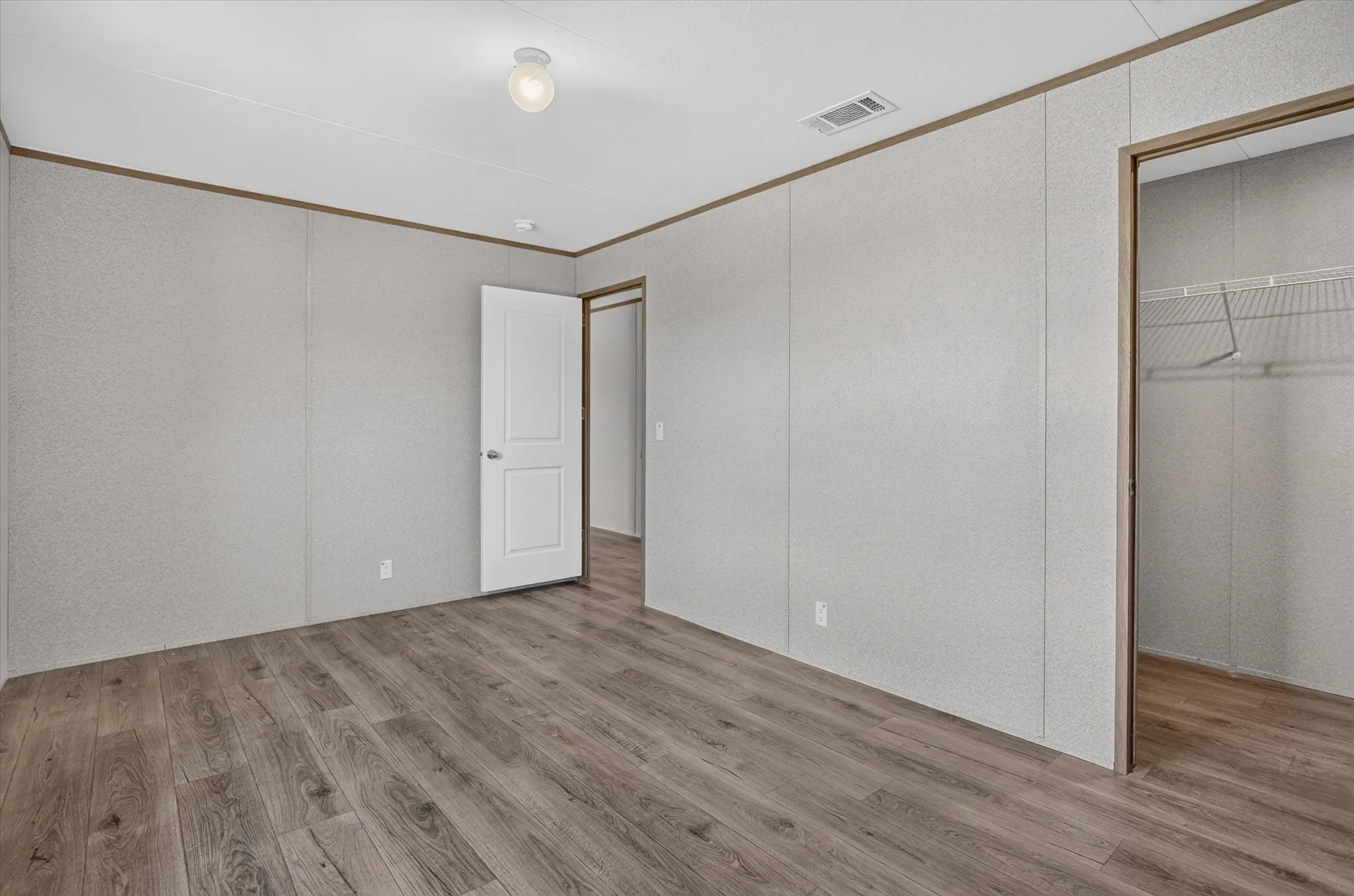 Spacious room featuring wood-finish flooring, light grey wall panels, a white flush-mount ceiling light fixture, and a closet with wire shelving