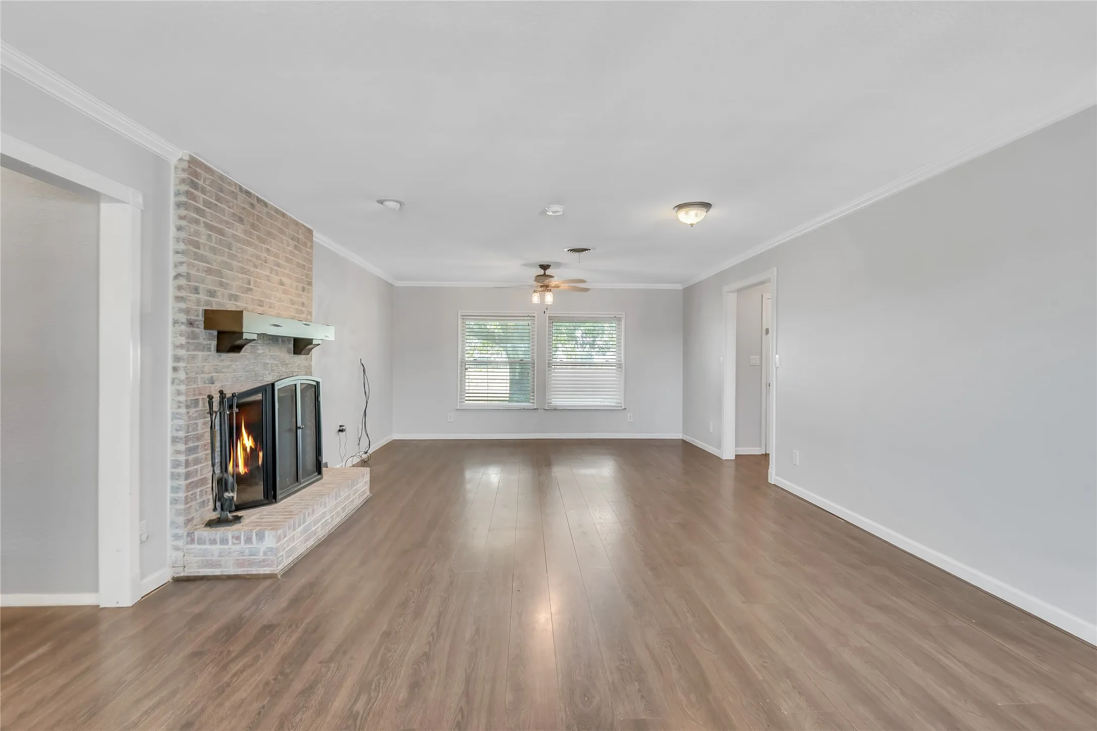 Wood-finish flooring throughout, brick fireplace with a mantle, dual windows with blinds, and ceiling fan with light fixture
