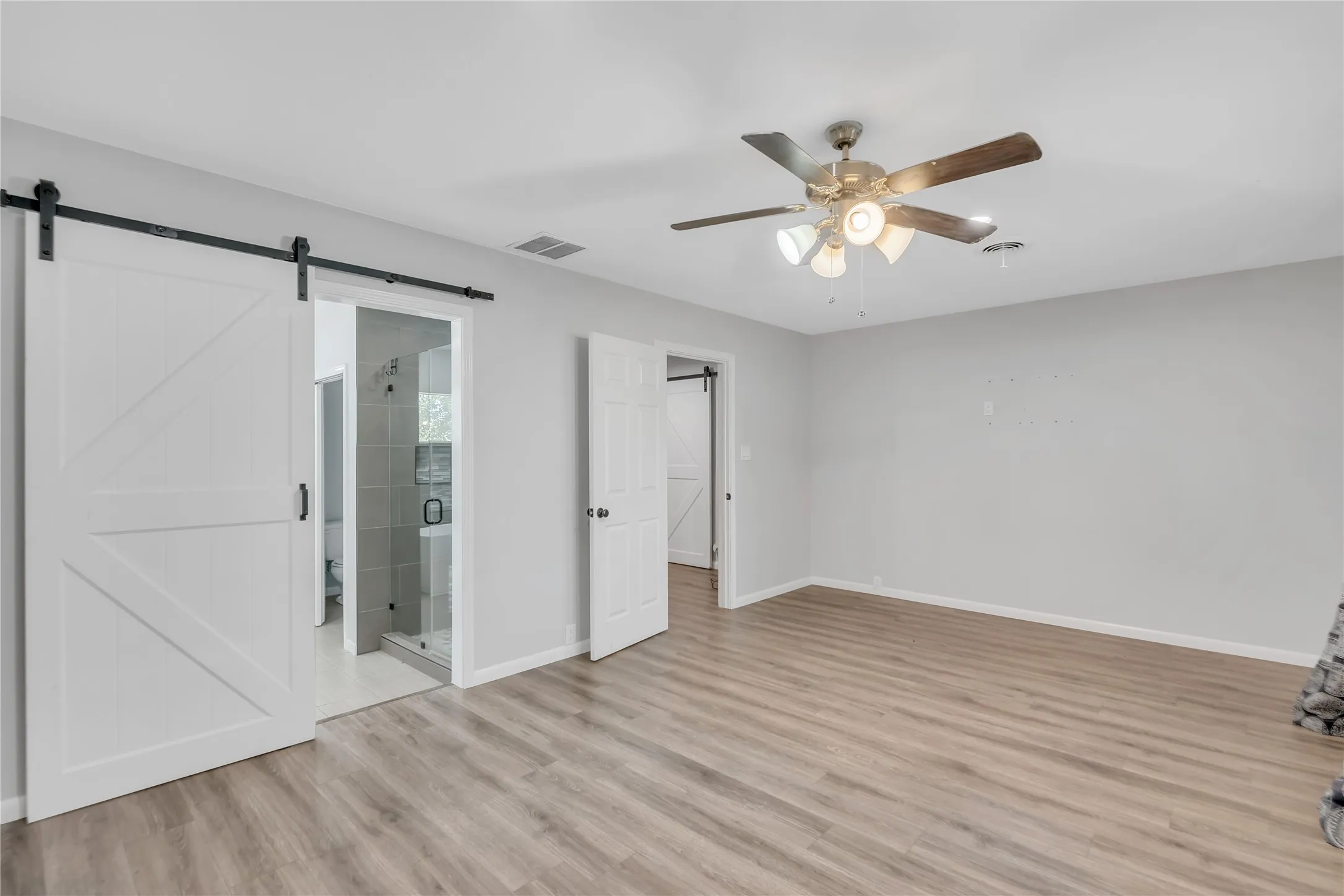 Wood-finish flooring, ceiling fan with integrated lighting, white barn door, and a partially visible shower enclosure with tiled walls