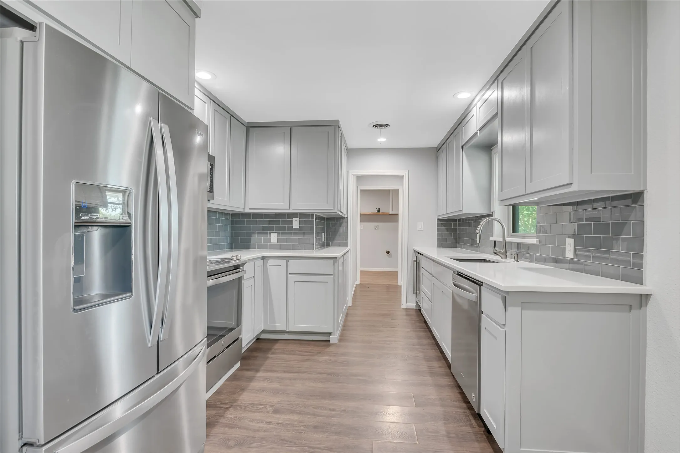 Galley kitchen with light gray cabinetry, stainless steel appliances, wood-finish flooring, white countertops, and a gray subway tile backsplash