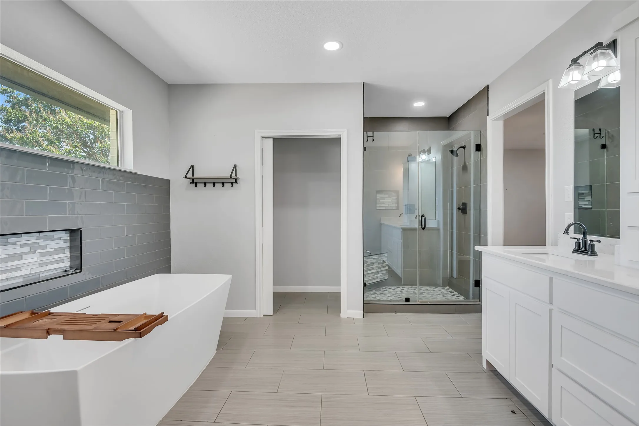 Freestanding soaking tub with tiled surround, glass-enclosed shower with tiled walls, dual vanity with undermount sinks, and light-toned tile flooring