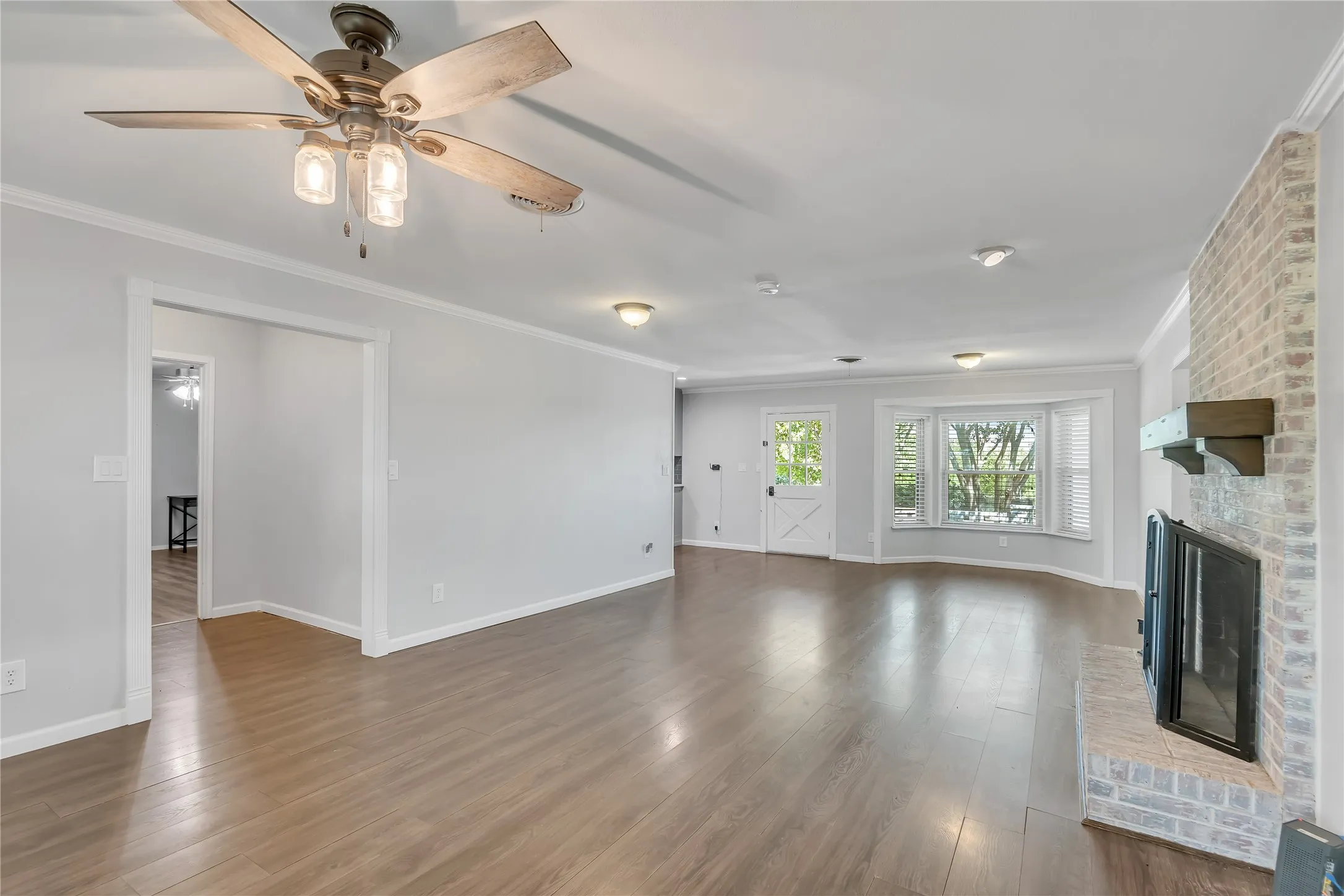 Wood-finish flooring, brick-faced fireplace with wood mantel, bay window, white interior door with crossbuck detail, and a ceiling fan with integrated lighting