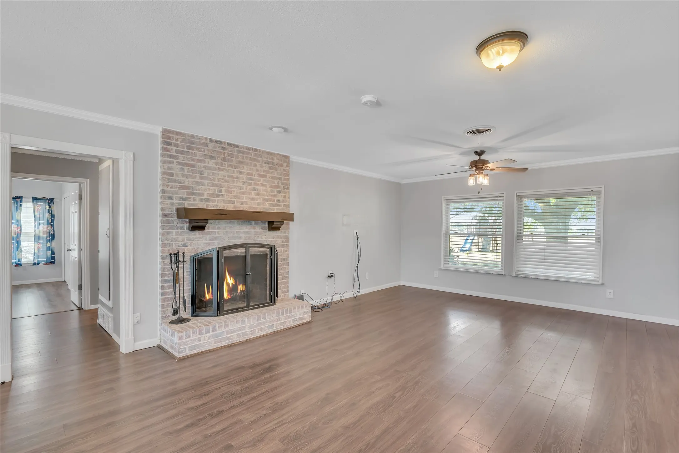 Spacious living area featuring wood-finish flooring, a brick-surround fireplace with wood mantel, two large windows with blinds, and a ceiling fan with integrated lighting