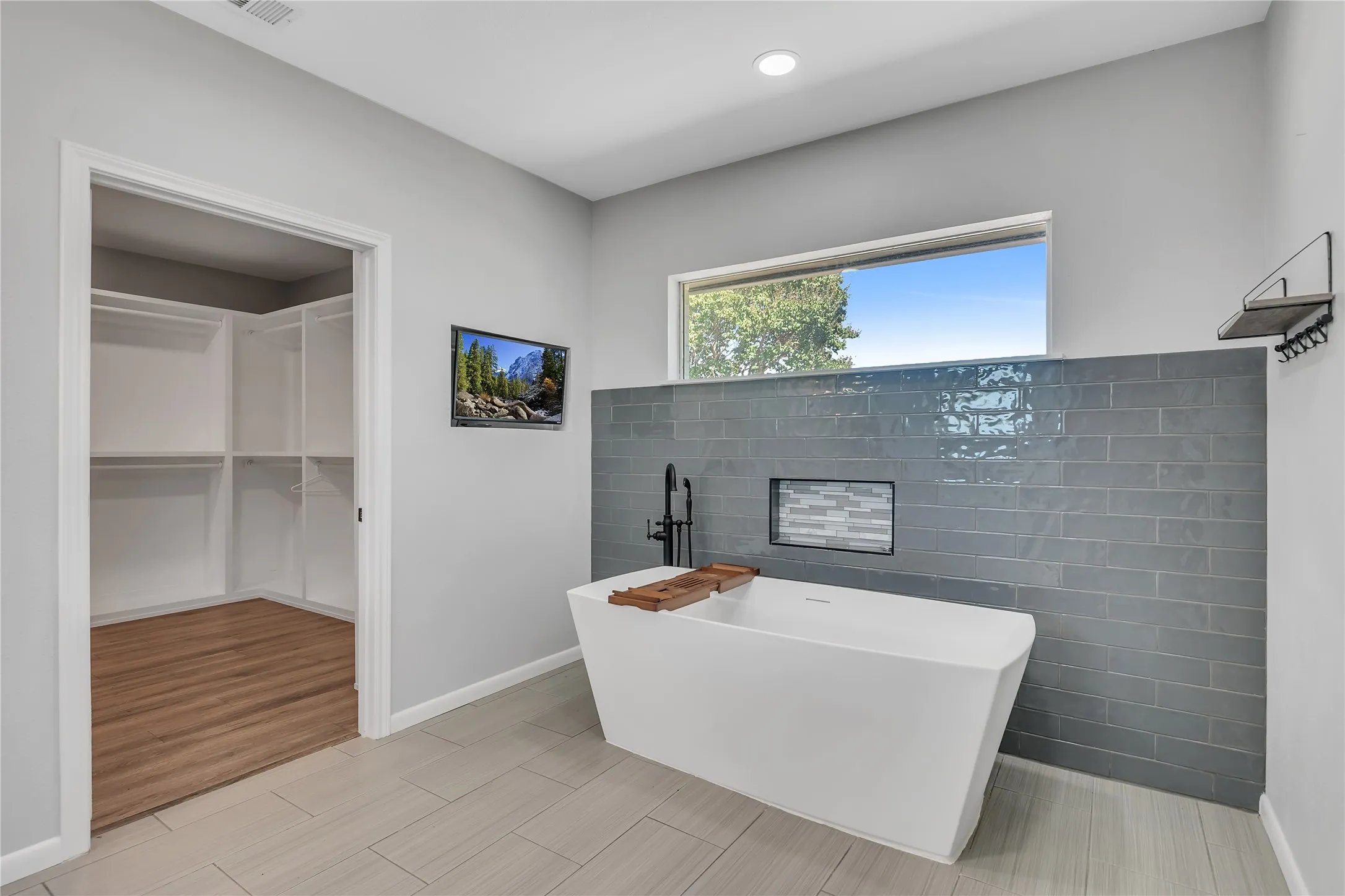 Freestanding soaking tub with a matte black fixture, accent tile wall with recessed shelf, and an adjacent walk-in closet with built-in shelving