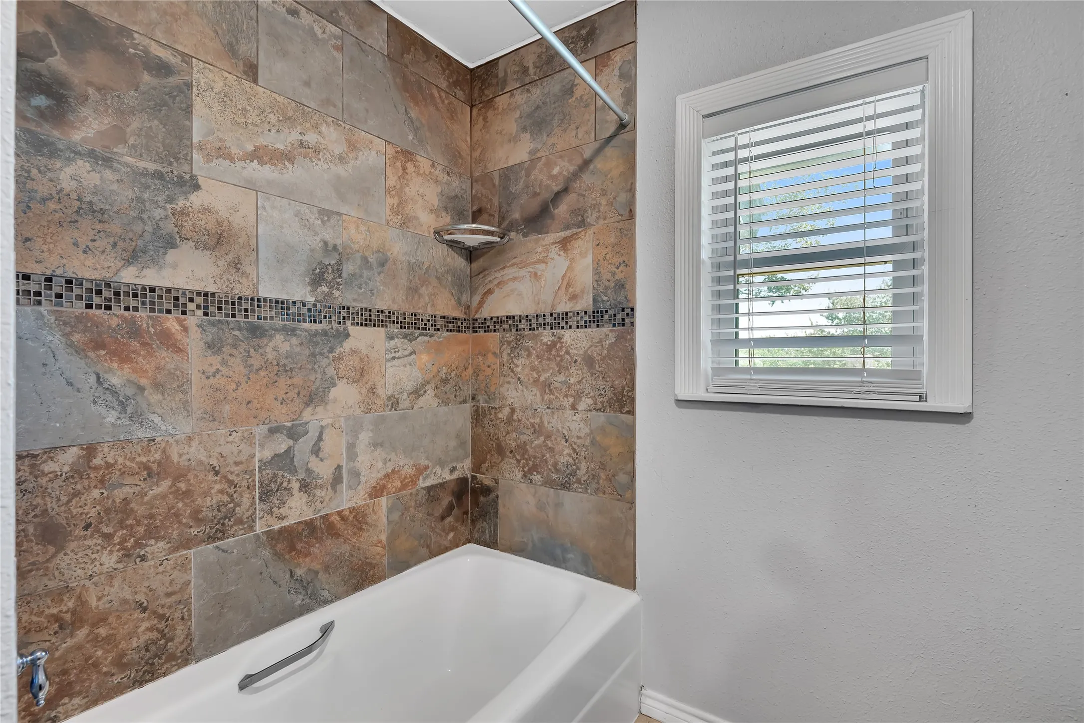 Bathtub with natural stone tile surround, decorative mosaic tile accent, chrome shower fixtures, and a window with blinds