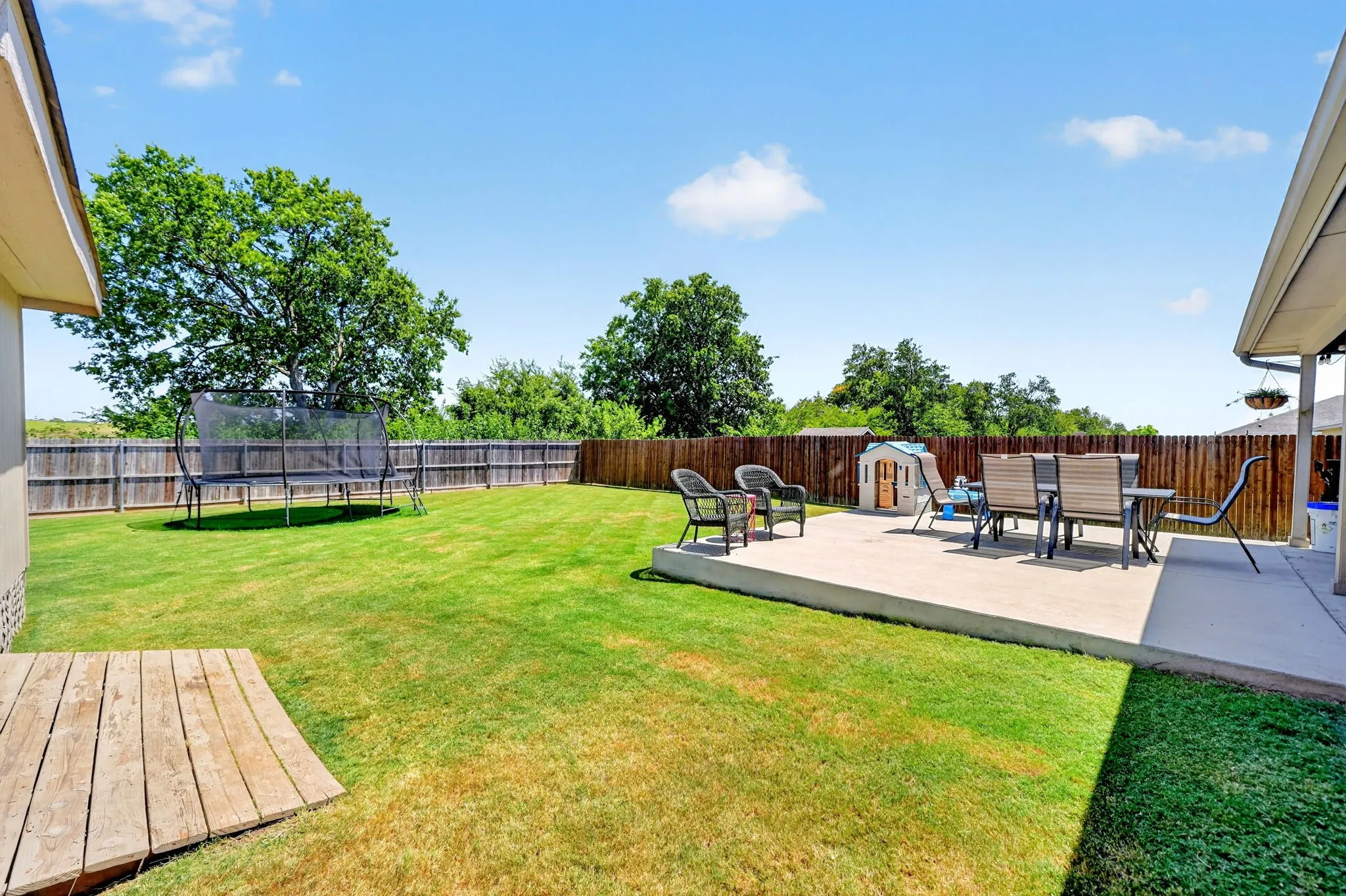 Expansive backyard featuring a manicured lawn, established trees, a substantial concrete patio, and a wood-panel fence