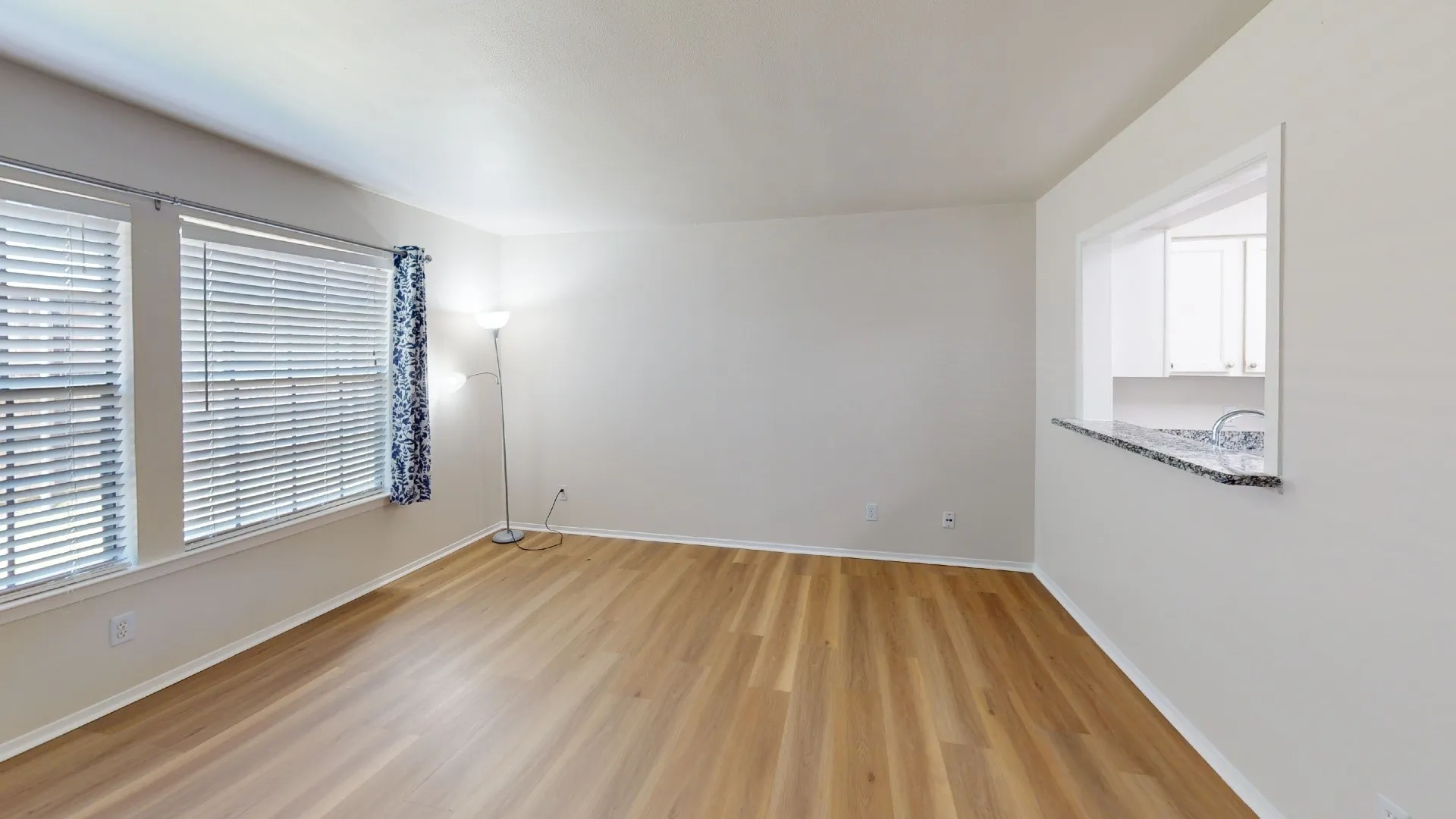 Expansive room featuring wood-finish flooring, two windows with horizontal blinds, and a pass-through opening with a granite-look countertop