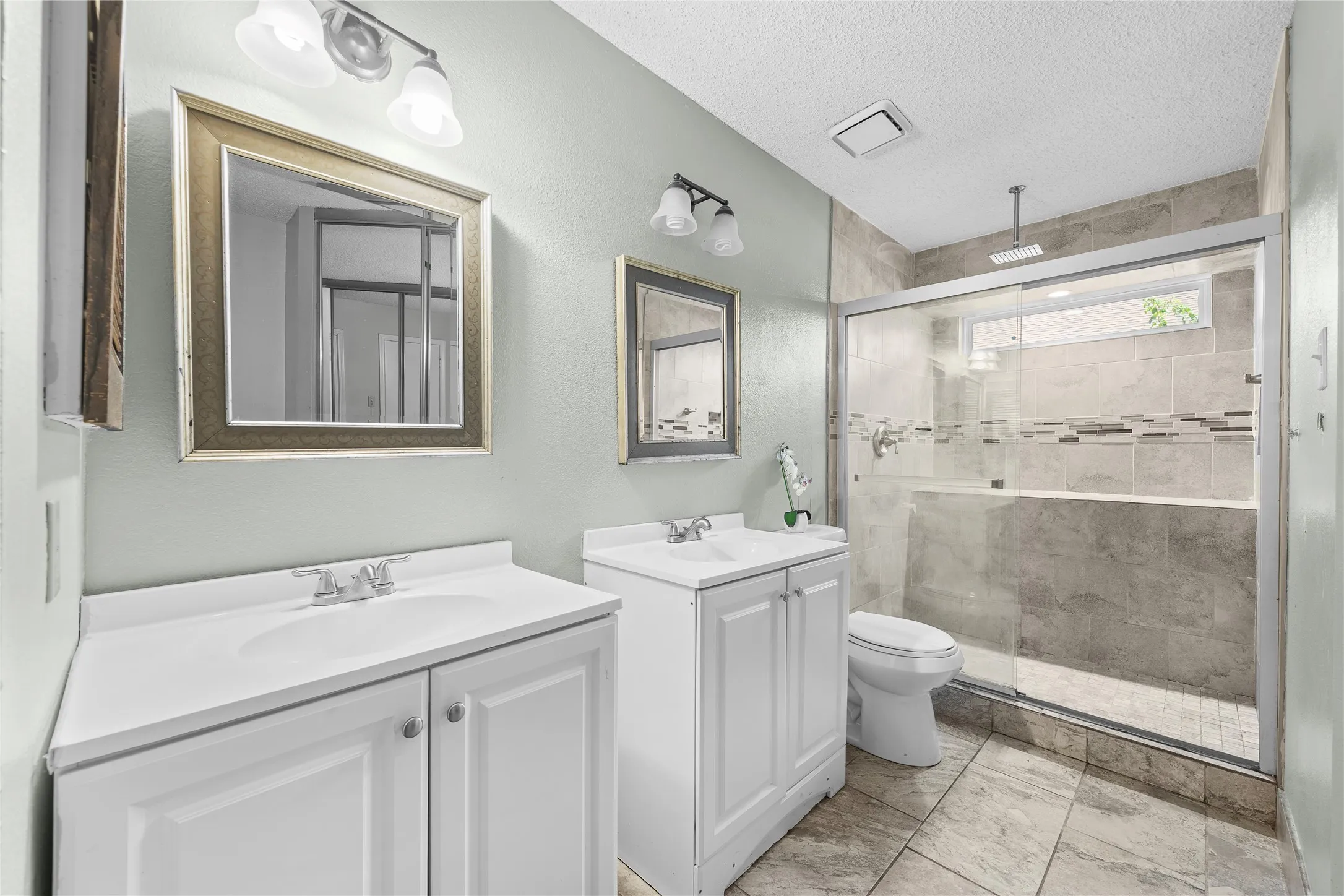 Dual vanity sinks with white cabinetry, framed mirrors, and a glass-enclosed shower featuring full-height tile and a horizontal decorative tile band