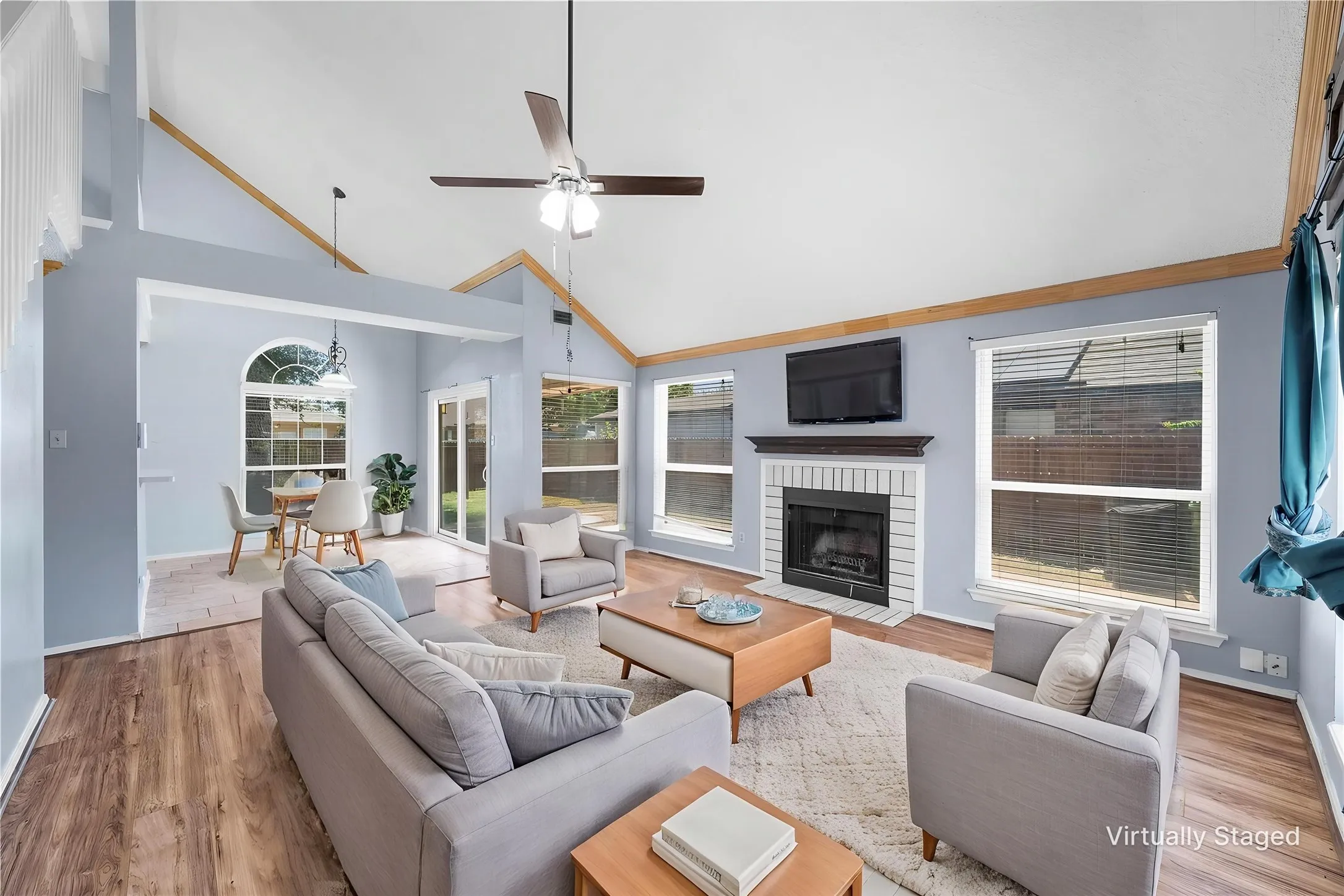 Vaulted ceilings, wood-finish flooring, white brick fireplace, multiple windows, and a ceiling fan