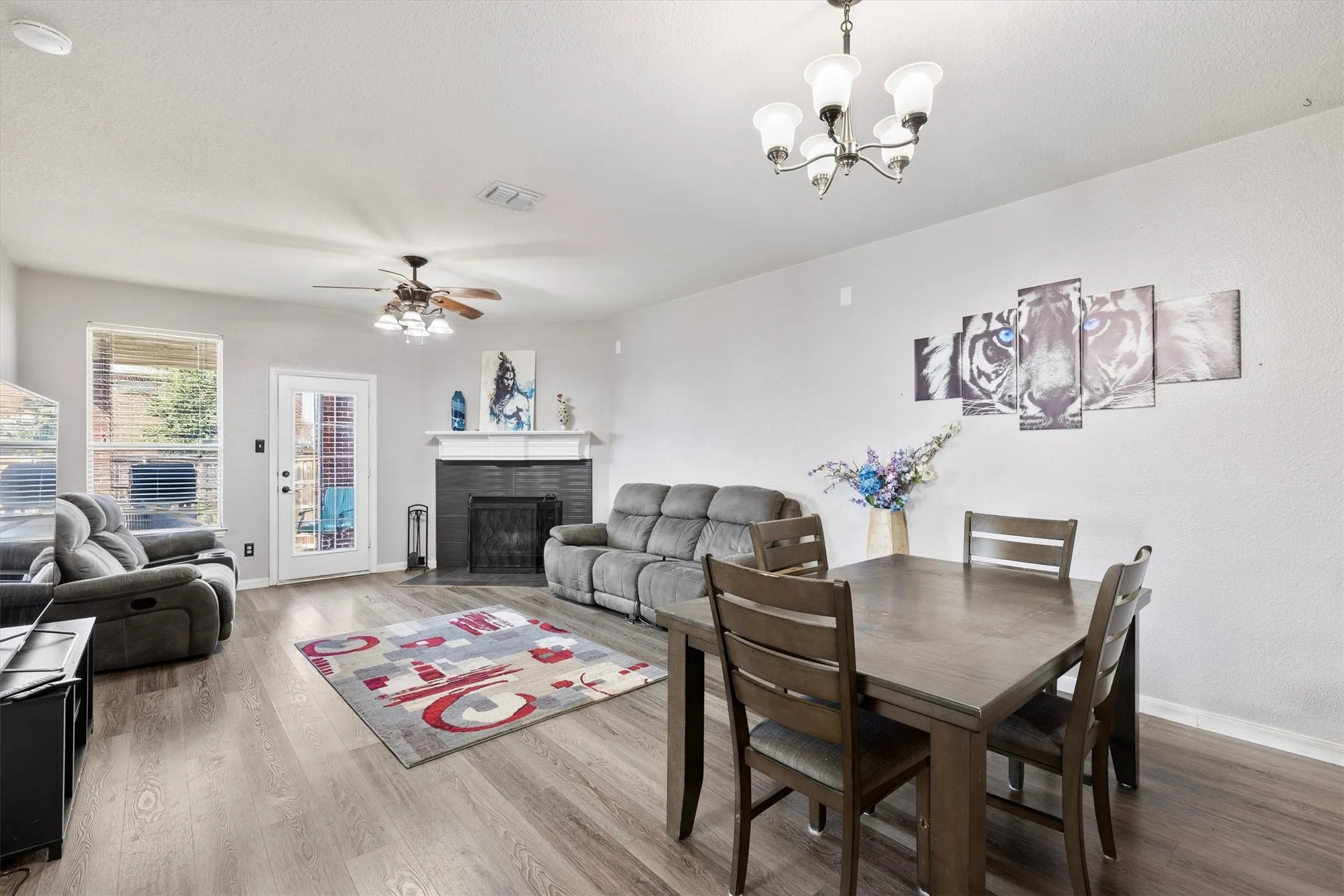 Wood-finish flooring, ceiling fan with integrated lighting, chandelier, white mantel, and a rear door with integrated blinds