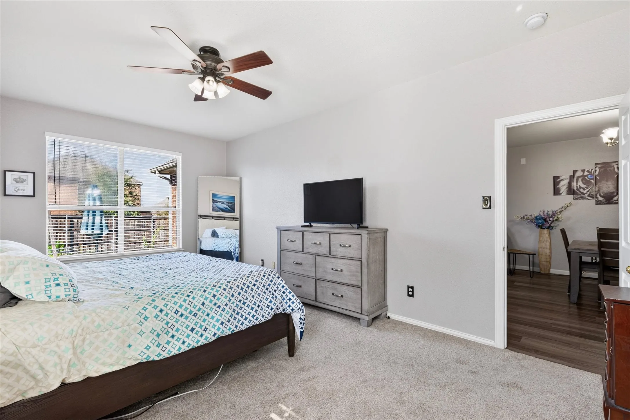 Light gray wall paint, textured carpet flooring, ceiling fan with integrated lighting, window with horizontal blinds, and a doorway leading to a room with wood-finish flooring