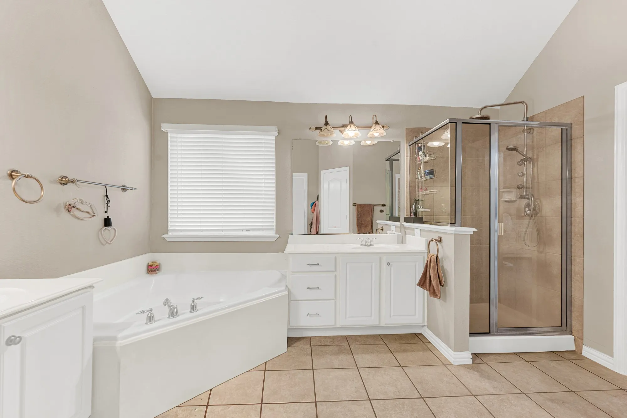Large corner soaking tub, glass-enclosed shower with multiple showerheads, dual vanity with white cabinetry, tile flooring, and vaulted ceiling