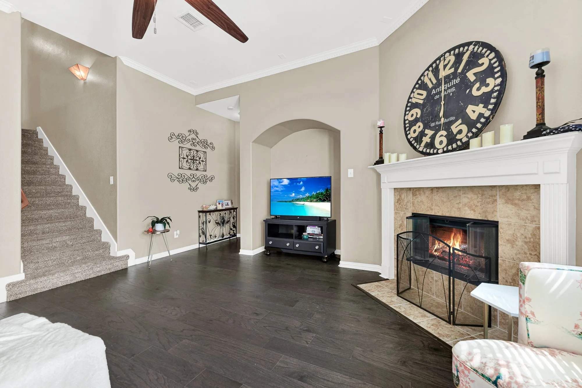 Wood-finish flooring, a ceiling fan, a fireplace with a tile surround and white mantel, and an arched entry