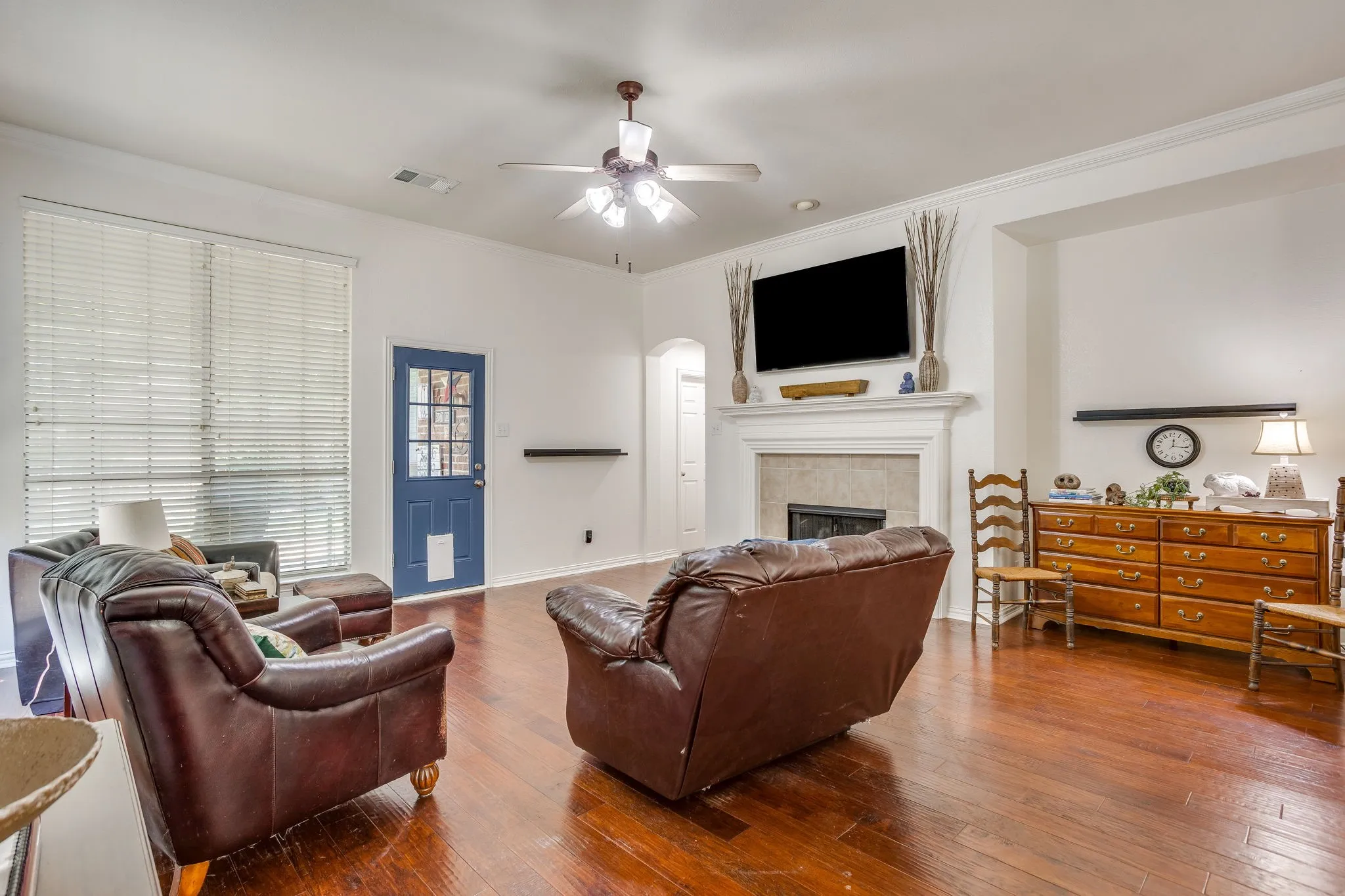 Hardwood flooring, white walls, ceiling fan with light fixture, fireplace with mantel, and a blue door with glass panels