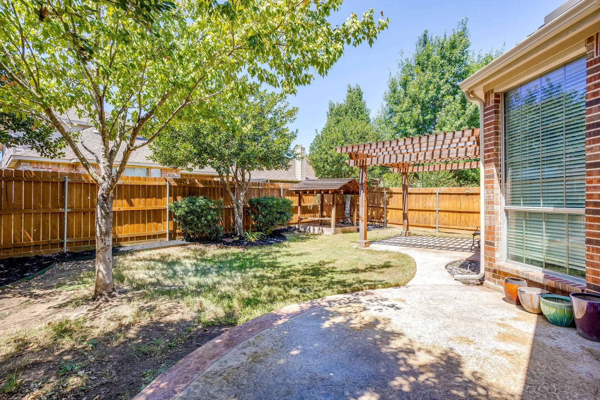 Spacious backyard oasis featuring mature shade trees, established landscaping with shrub borders, wooden privacy fencing, concrete patio area, and brick home exterior with large window installations
