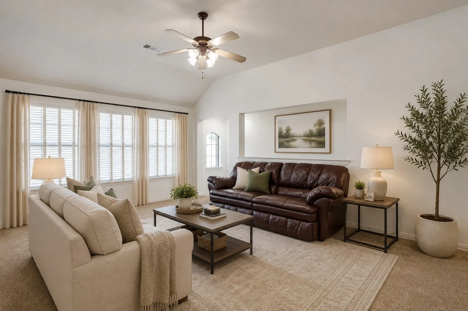 Bright living room featuring multiple large windows with plantation shutters and natural light, ceiling fan with integrated lighting, neutral wood-finish flooring, area rug, and mixed seating arrangements with side tables and table lamps throughout the space. Photo virtually staged using AI.