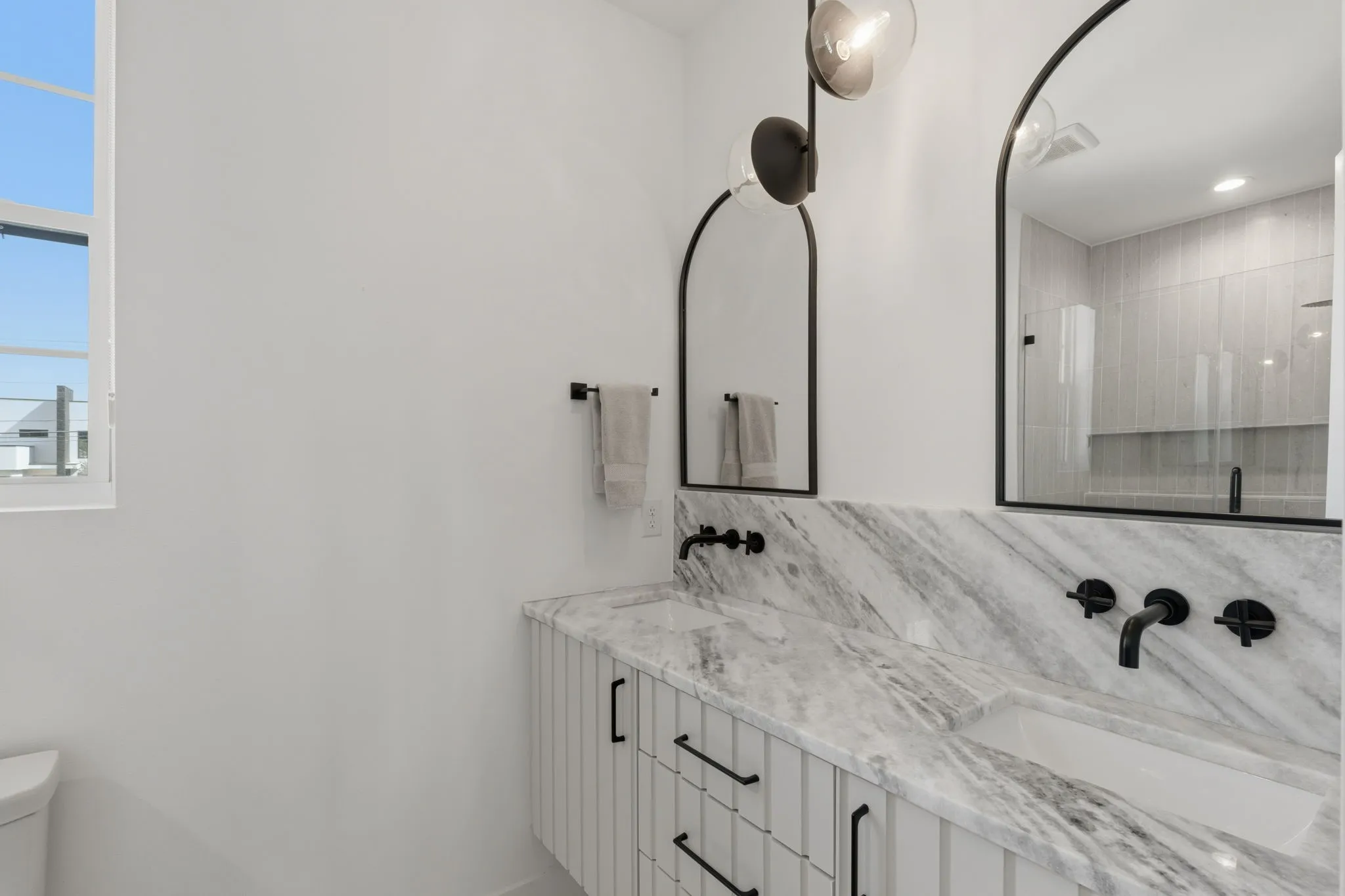 Dual vanity with stone countertop, integrated sinks, black wall-mounted faucets, and arched mirrors