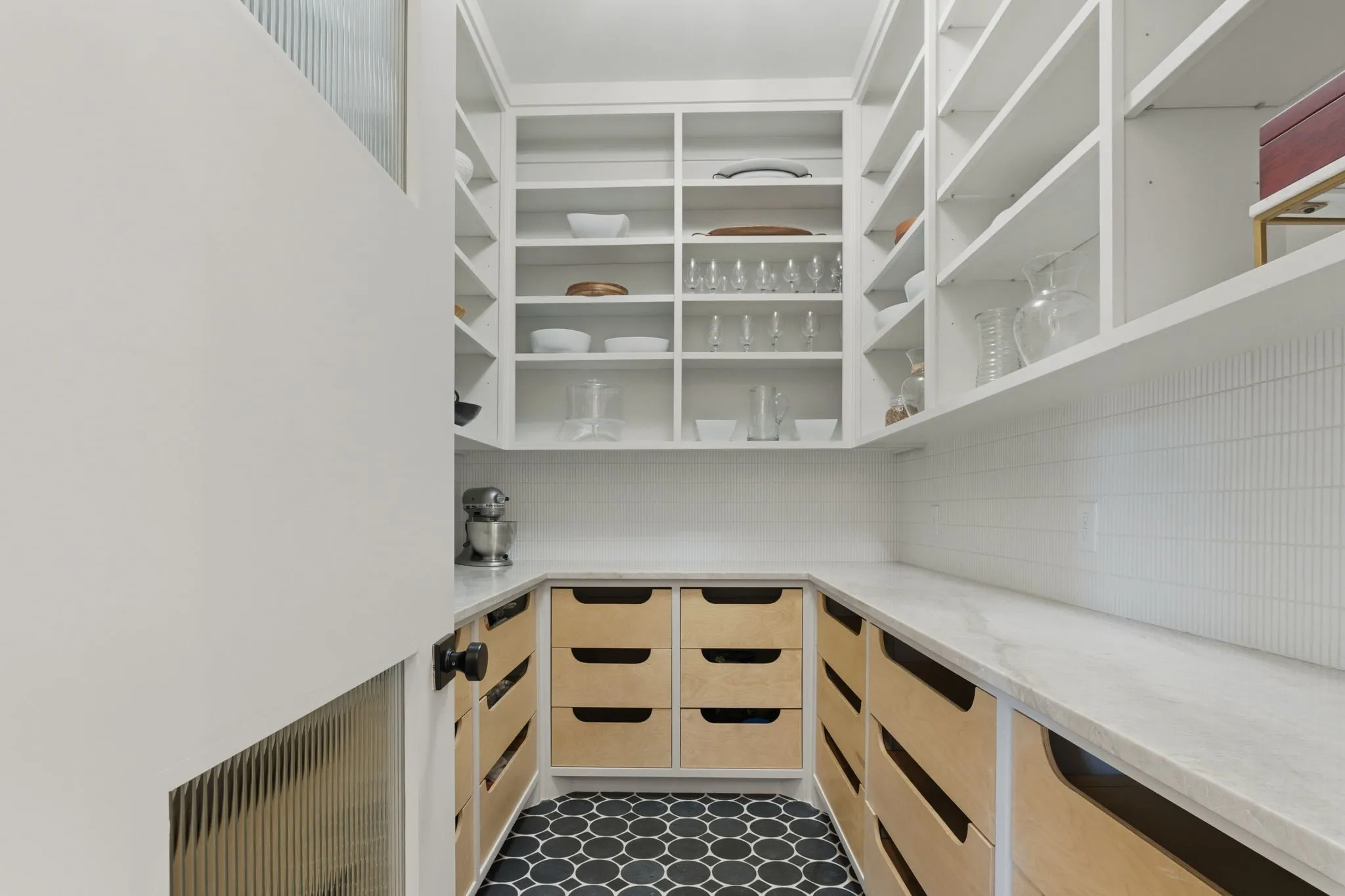 Sophisticated walk-in pantry featuring custom wood-finish cabinetry with pull-out drawer organization, white open shelving with integrated lighting, marble-look countertops, and geometric patterned flooring