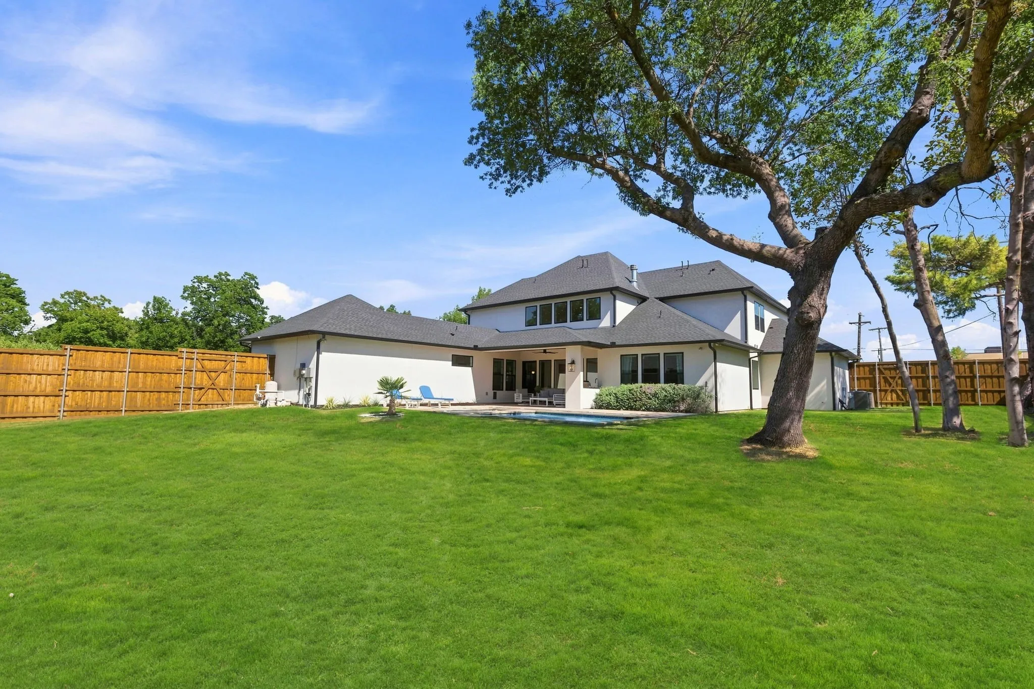 Expansive green lawn with mature trees, a privacy fence, and a multi-level white exterior residence featuring numerous windows