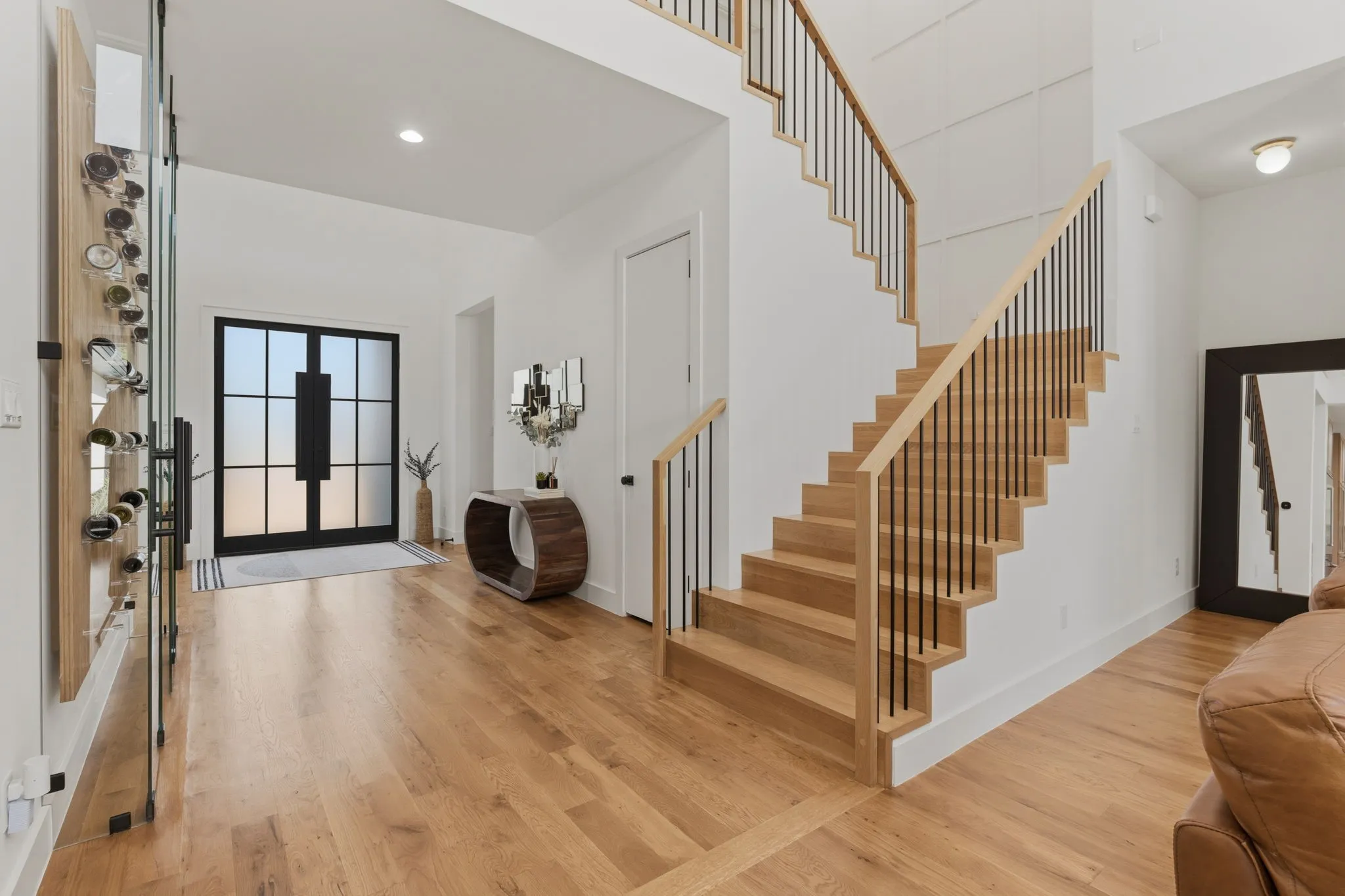 Wide plank wood-finish flooring, a contemporary staircase with wood treads and black metal balusters, and a double entry door with frosted glass and black framing