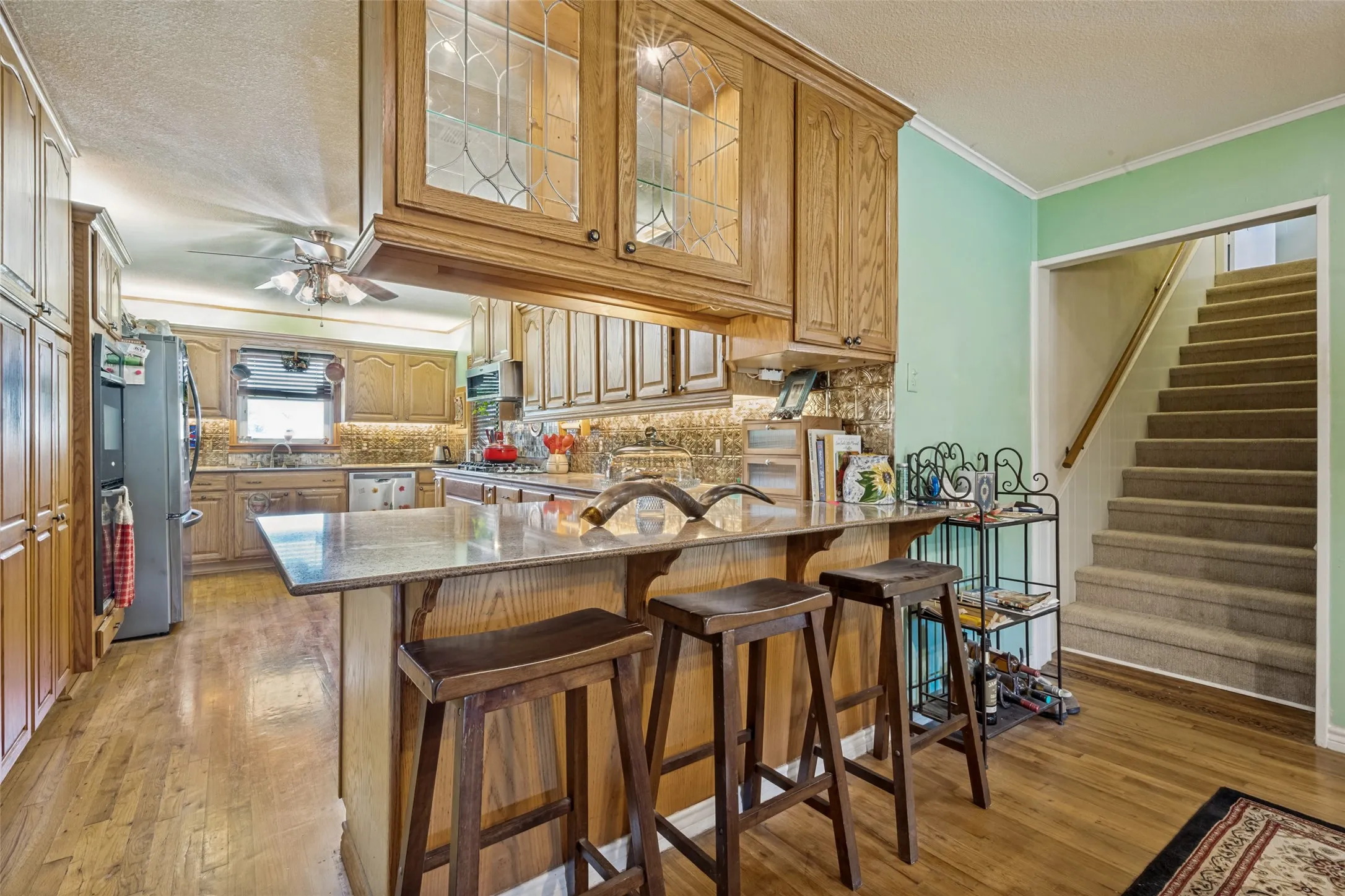Kitchen featuring wood-finish flooring, built-in wooden cabinetry, a granite-finish island with seating, a tile backsplash, and stainless steel appliances
