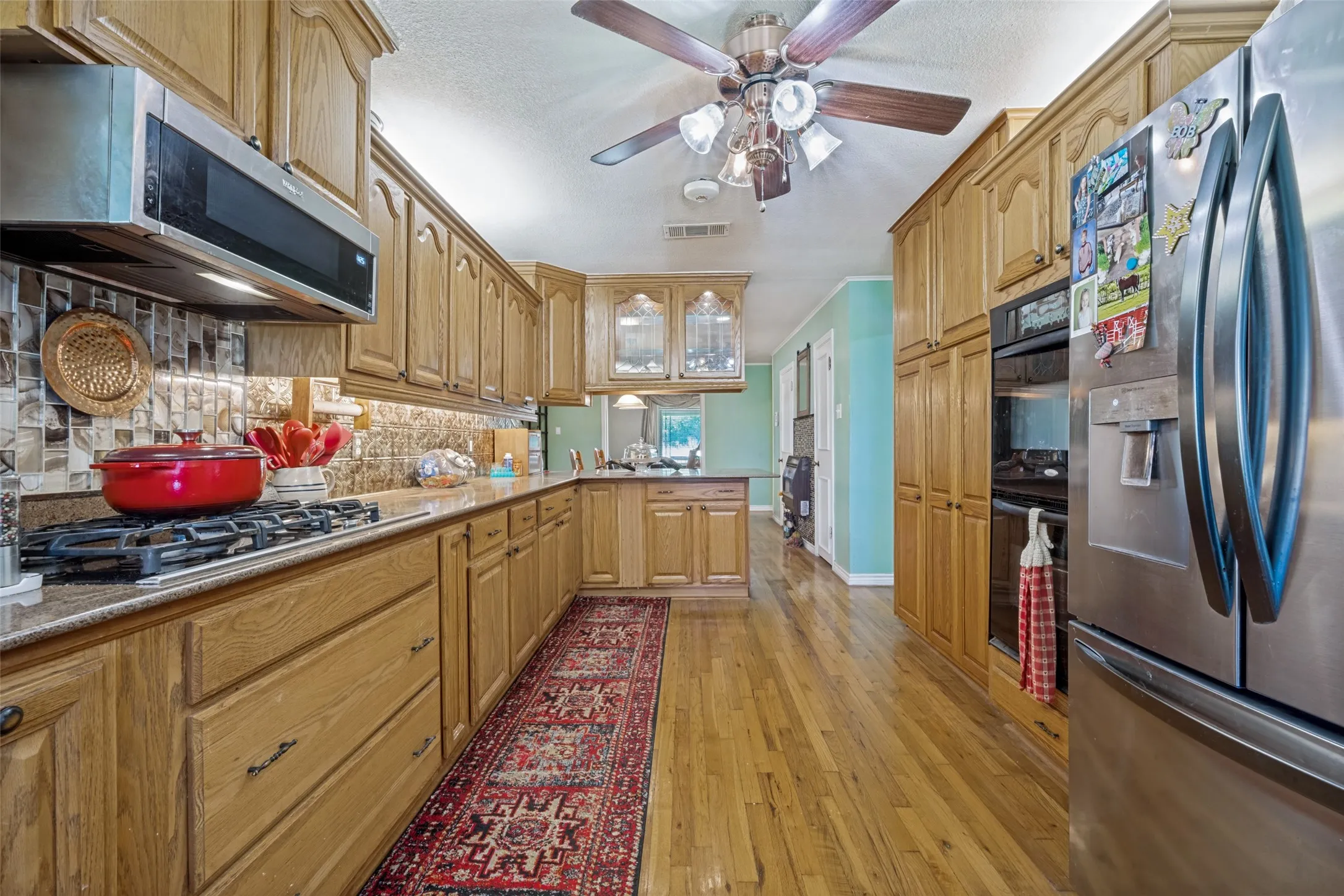 Kitchen featuring wood-finish flooring, extensive wood cabinetry, stainless steel appliances, a tile backsplash, and a ceiling fan
