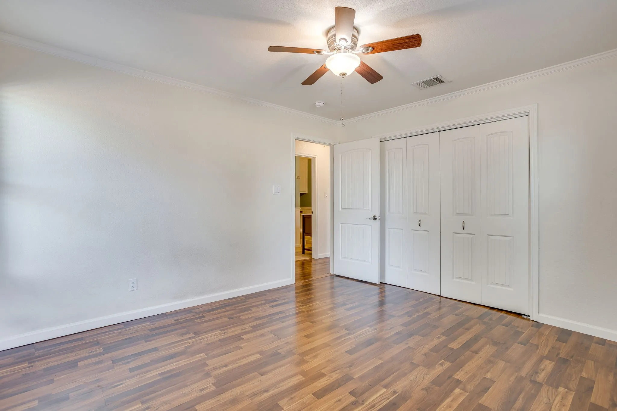 Wood laminate flooring, white walls, ceiling fan, crown molding, and bi-fold closet doors