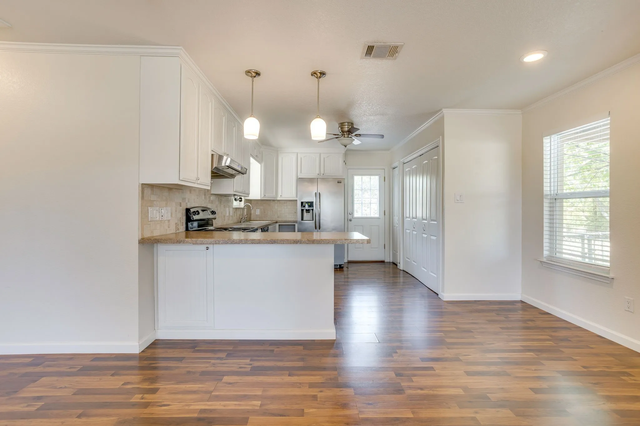 Open-concept living area featuring wood laminate flooring, white cabinetry, a kitchen island with seating, pendant lighting, and a window with blinds