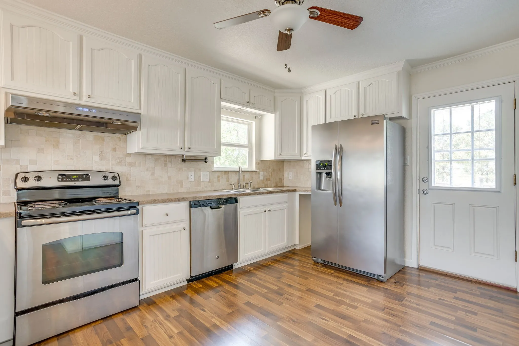 White cabinetry with crown molding, stainless steel appliances, patterned tile backsplash, wood laminate flooring, and a ceiling fan