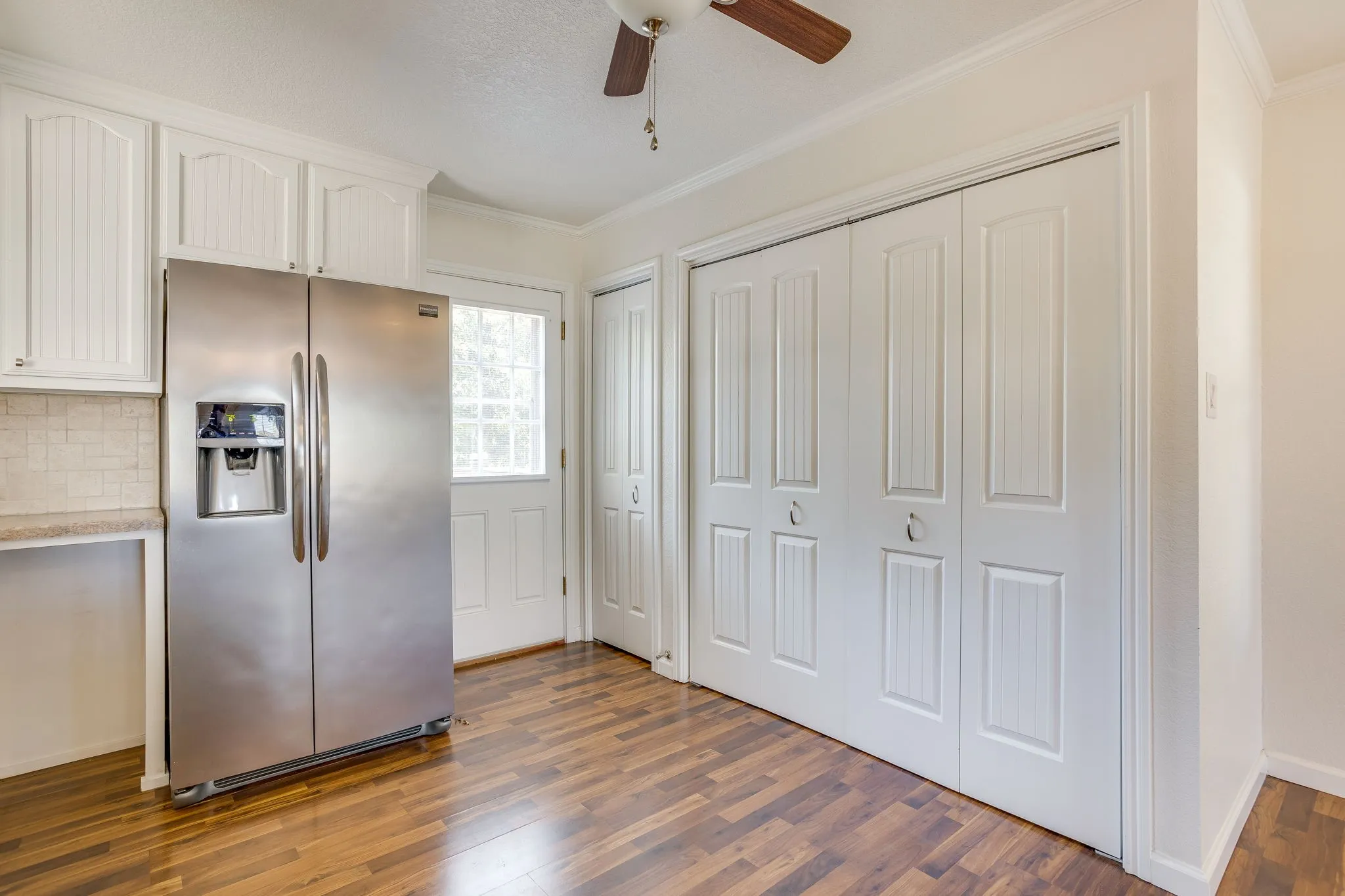 White cabinetry with beaded paneling, stainless steel refrigerator, light-toned tiled backsplash, wood laminate flooring, and white bi-fold closet doors